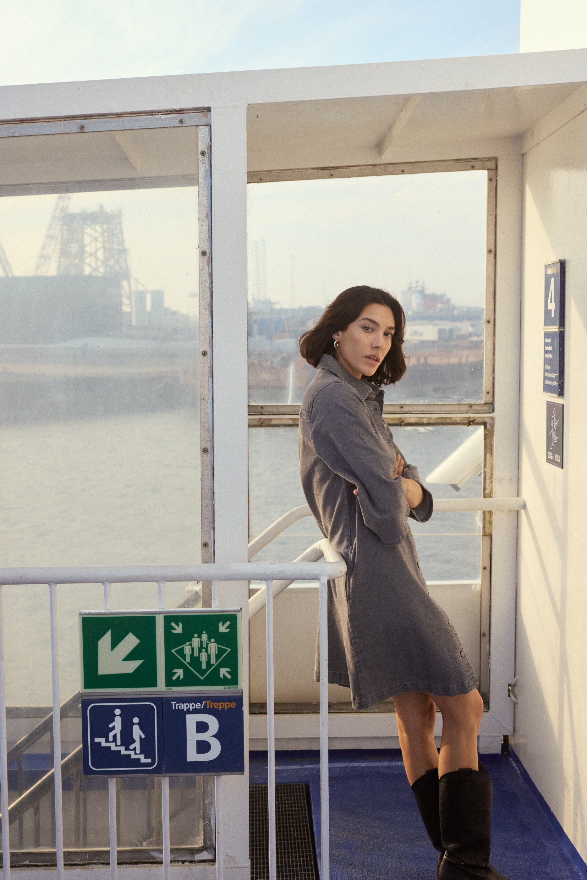 Woman in a grey dress and black boots leans on a boat deck railing, industrial backdrop.