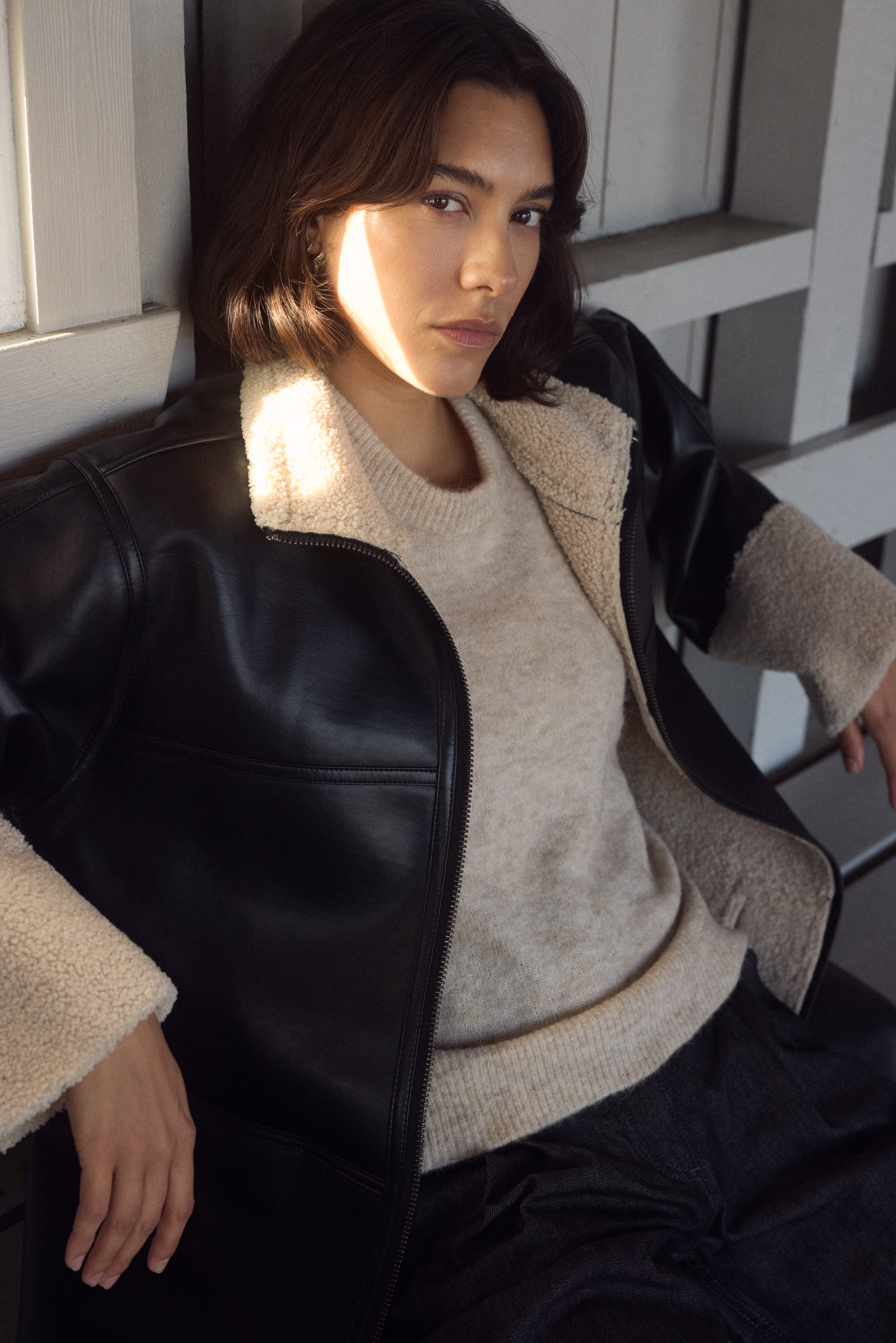Portrait of a woman in a black sherpa-lined jacket and beige sweater, lit by sunlight.
