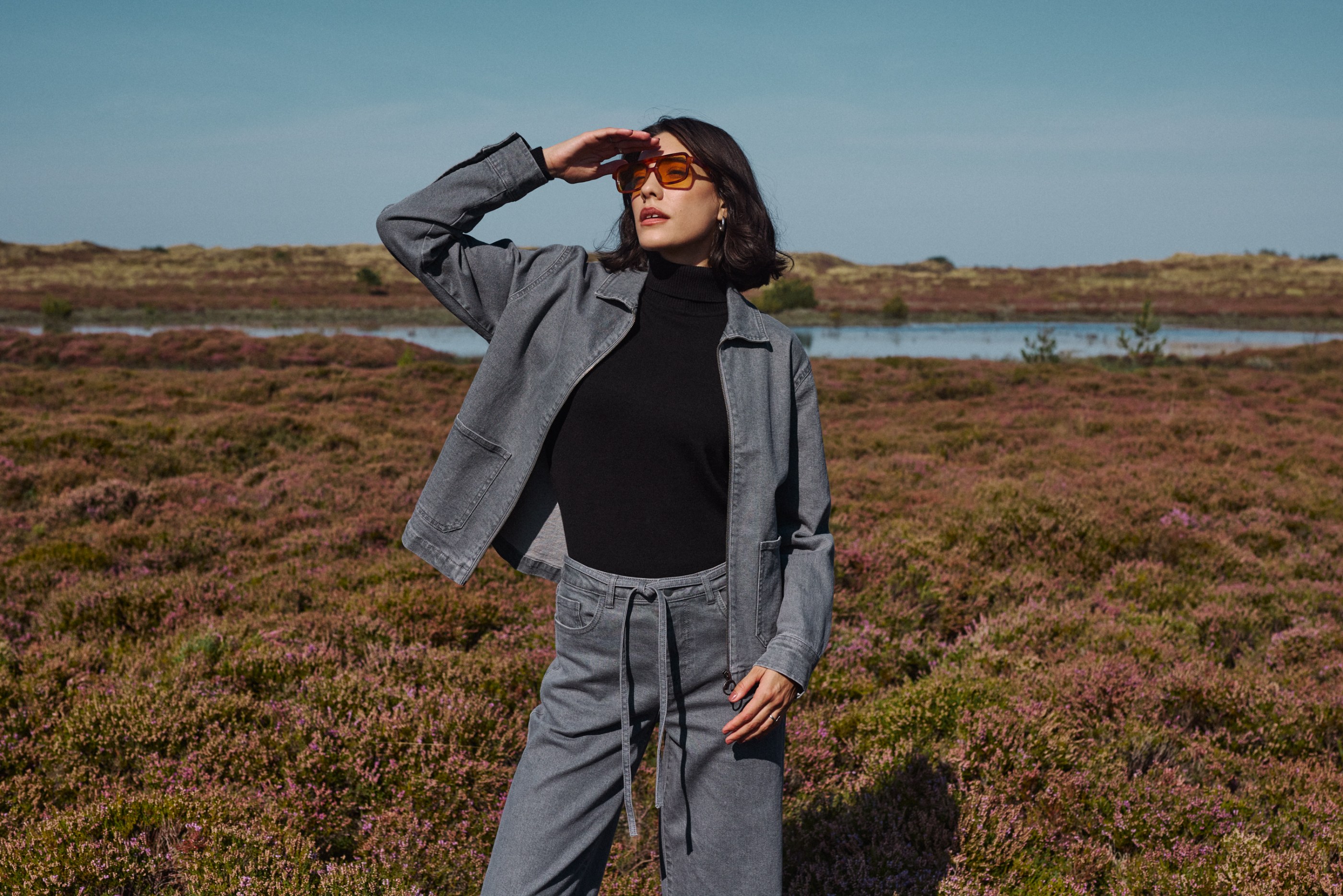 Woman in grey denim jacket and pants, orange sunglasses, shielding eyes in a blooming heather field.