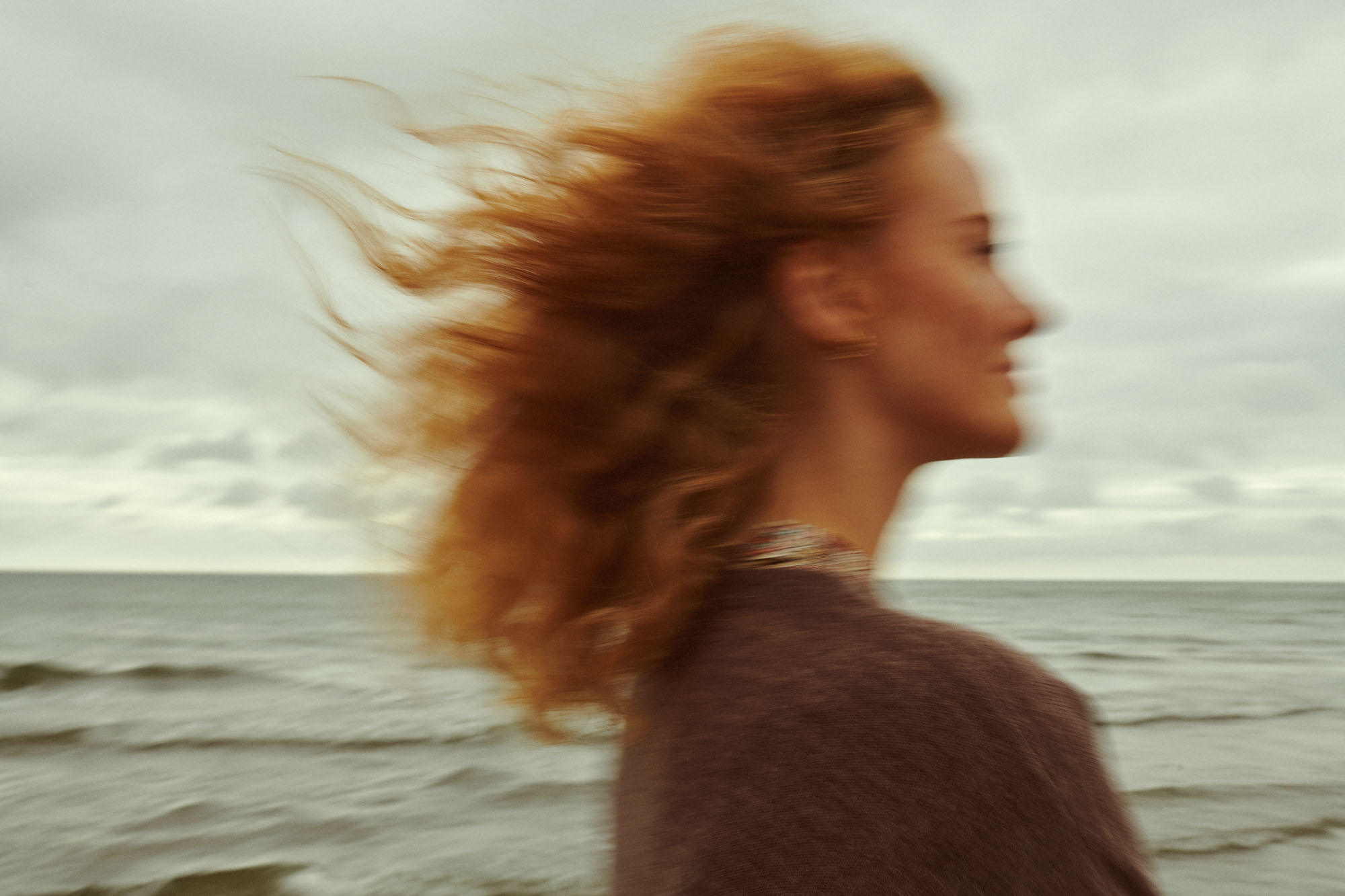 Blurred profile of a redhead woman with windblown hair by a cloudy ocean.