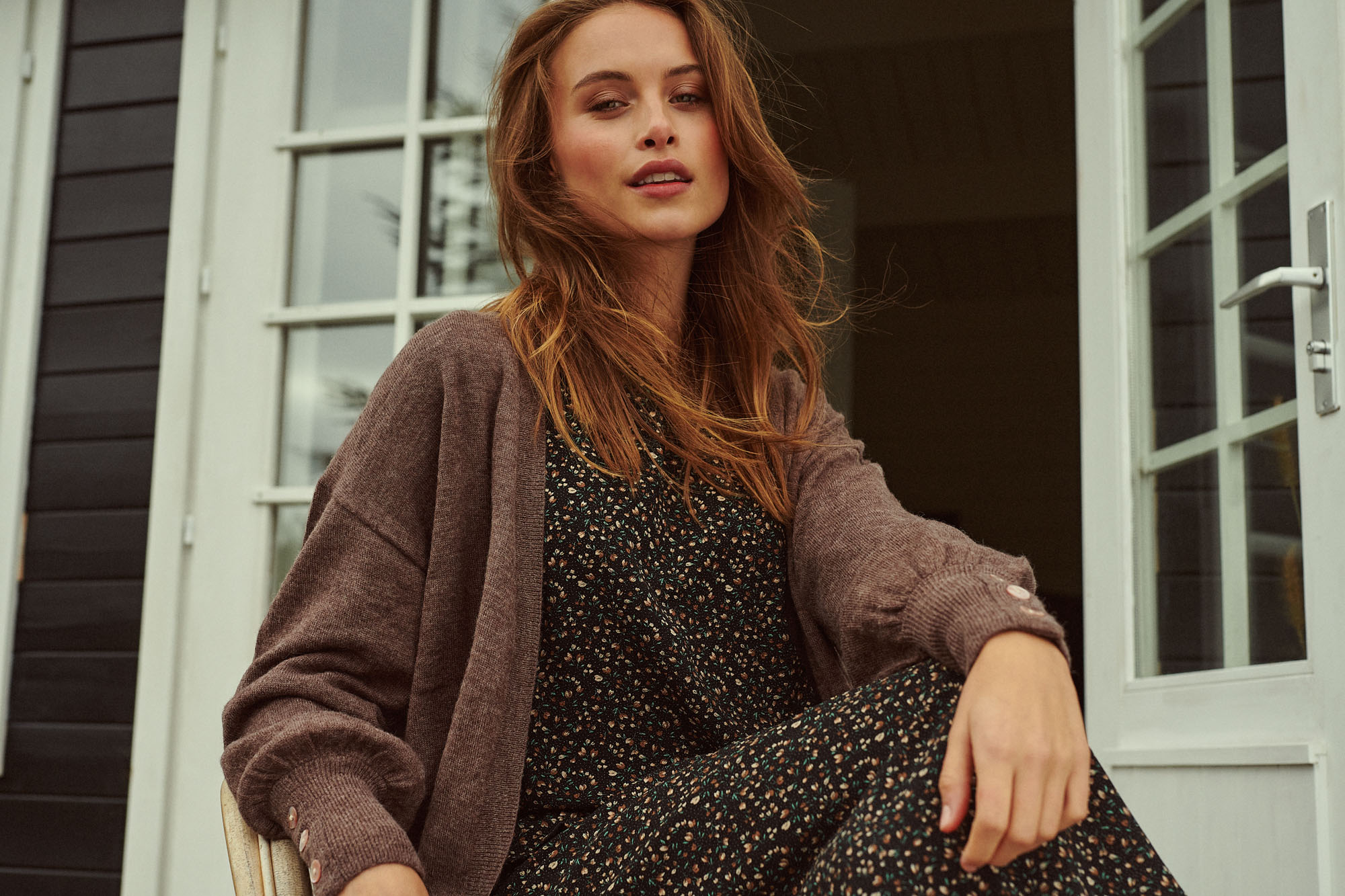 Woman in a brown cardigan and floral dress by a white window looks directly at the camera.