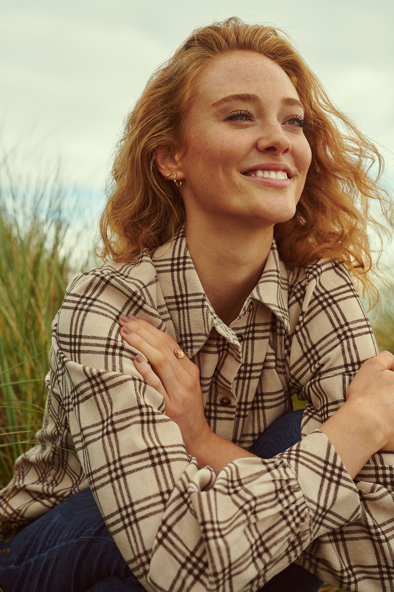 Smiling red-haired woman with freckles wearing a plaid shirt, sitting in tall grass outdoors.