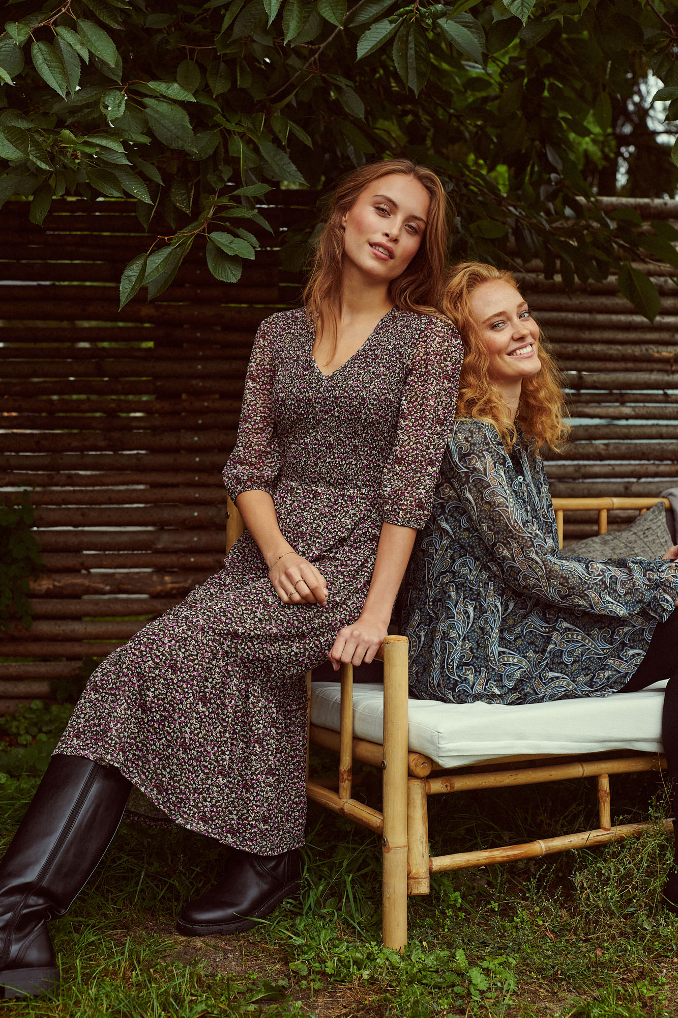 Two smiling women in floral and paisley dresses sitting on a bamboo bench outdoors.