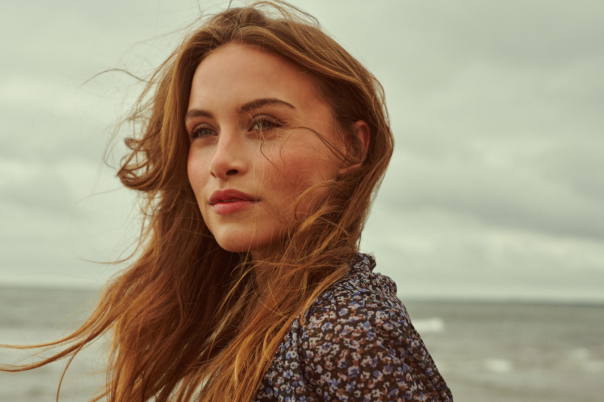 Woman with windblown auburn hair and floral dress looking thoughtfully at the cloudy sea.