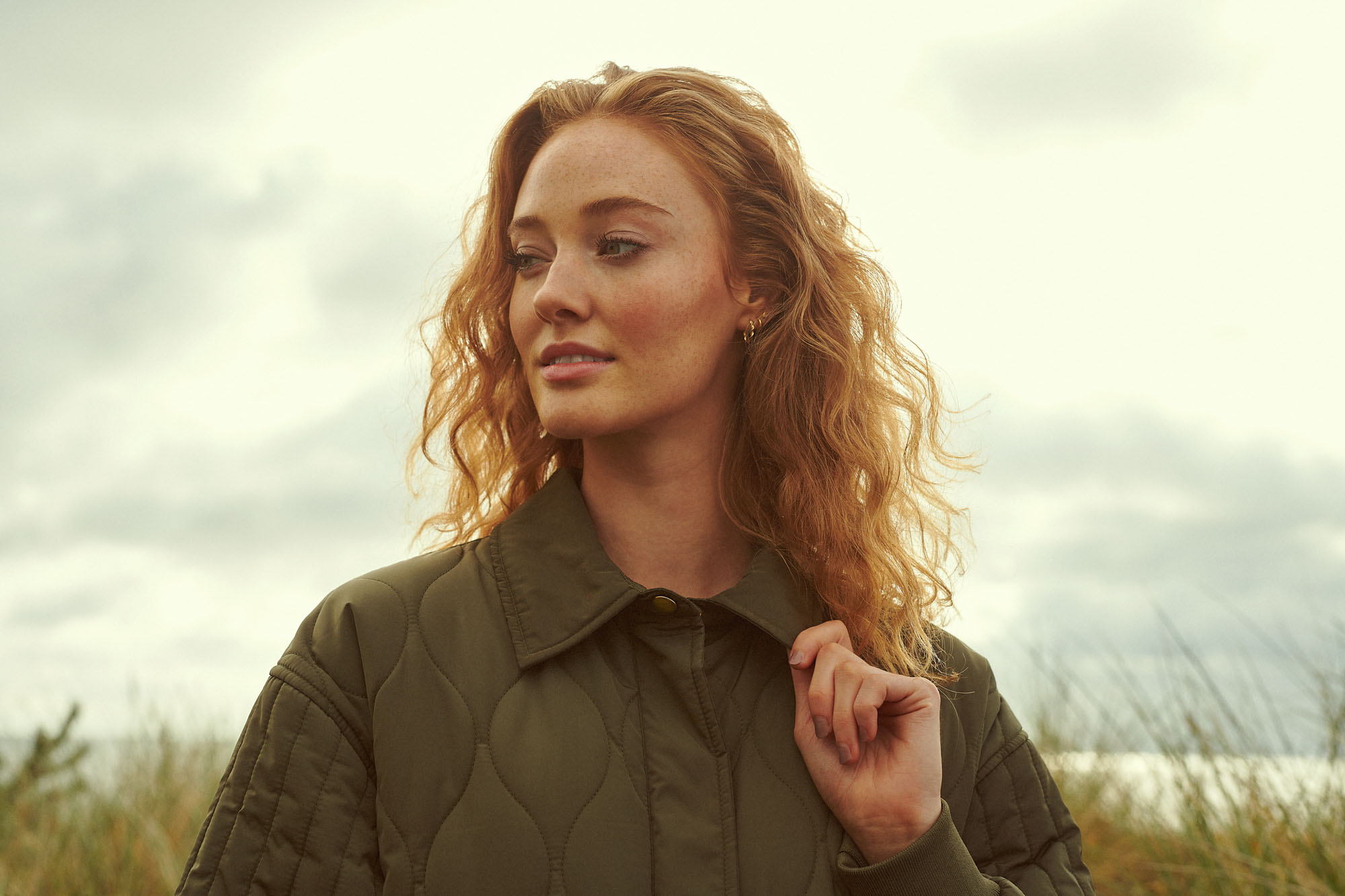 Red-haired woman in a green quilted jacket, looking pensively left against a cloudy sky.