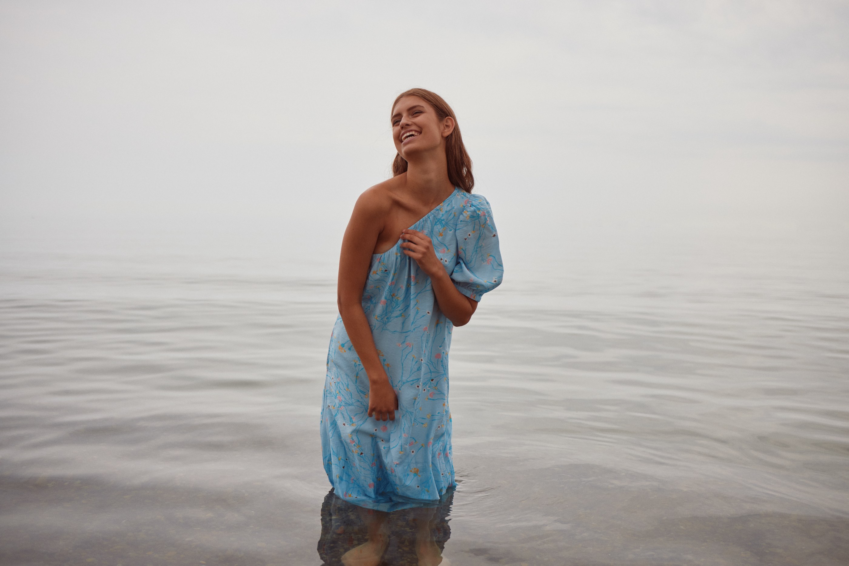 Young woman in a blue floral dress stands in water, smiling brightly under an overcast sky.