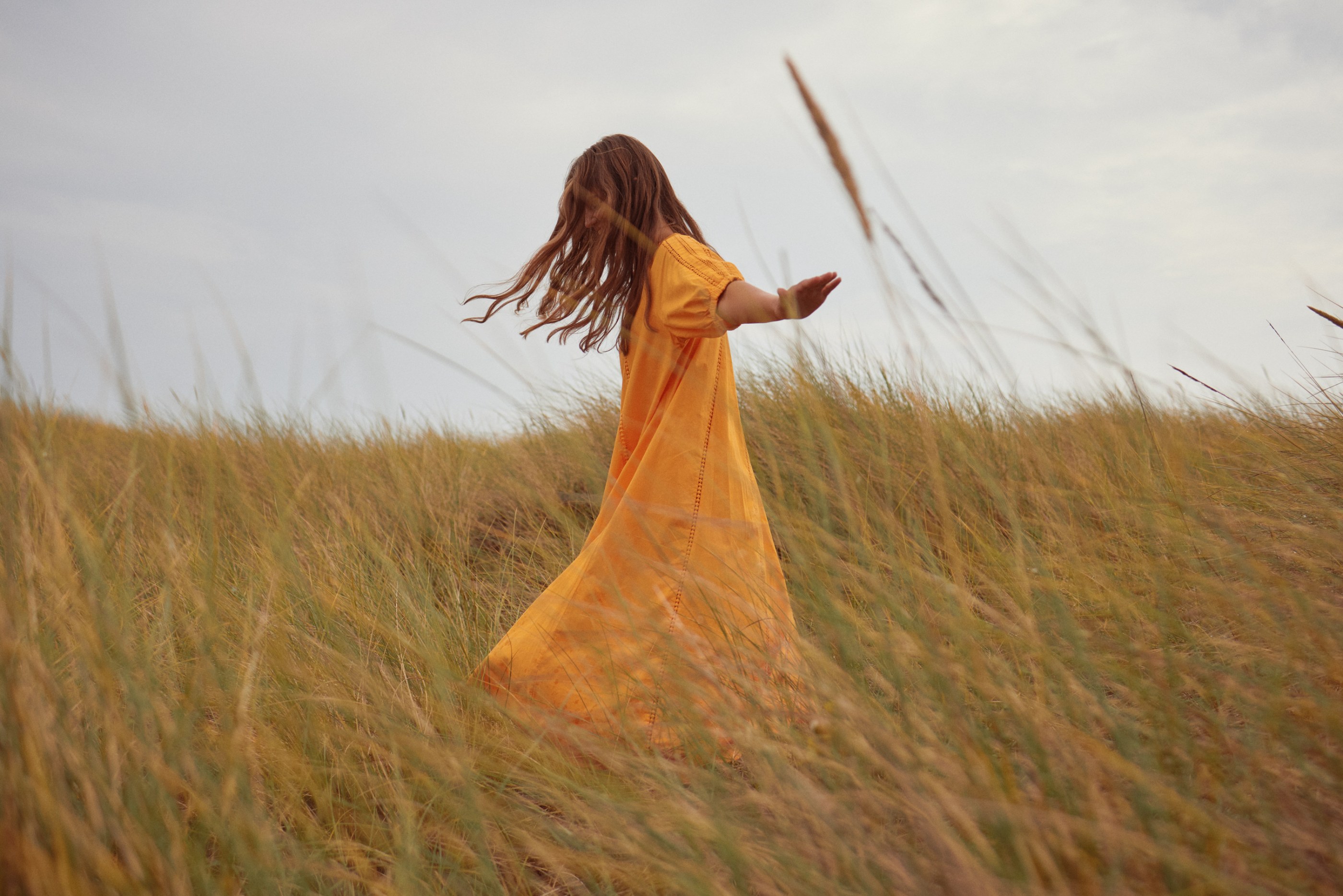 Person in a flowing yellow dress dancing through tall golden grass under an overcast sky.