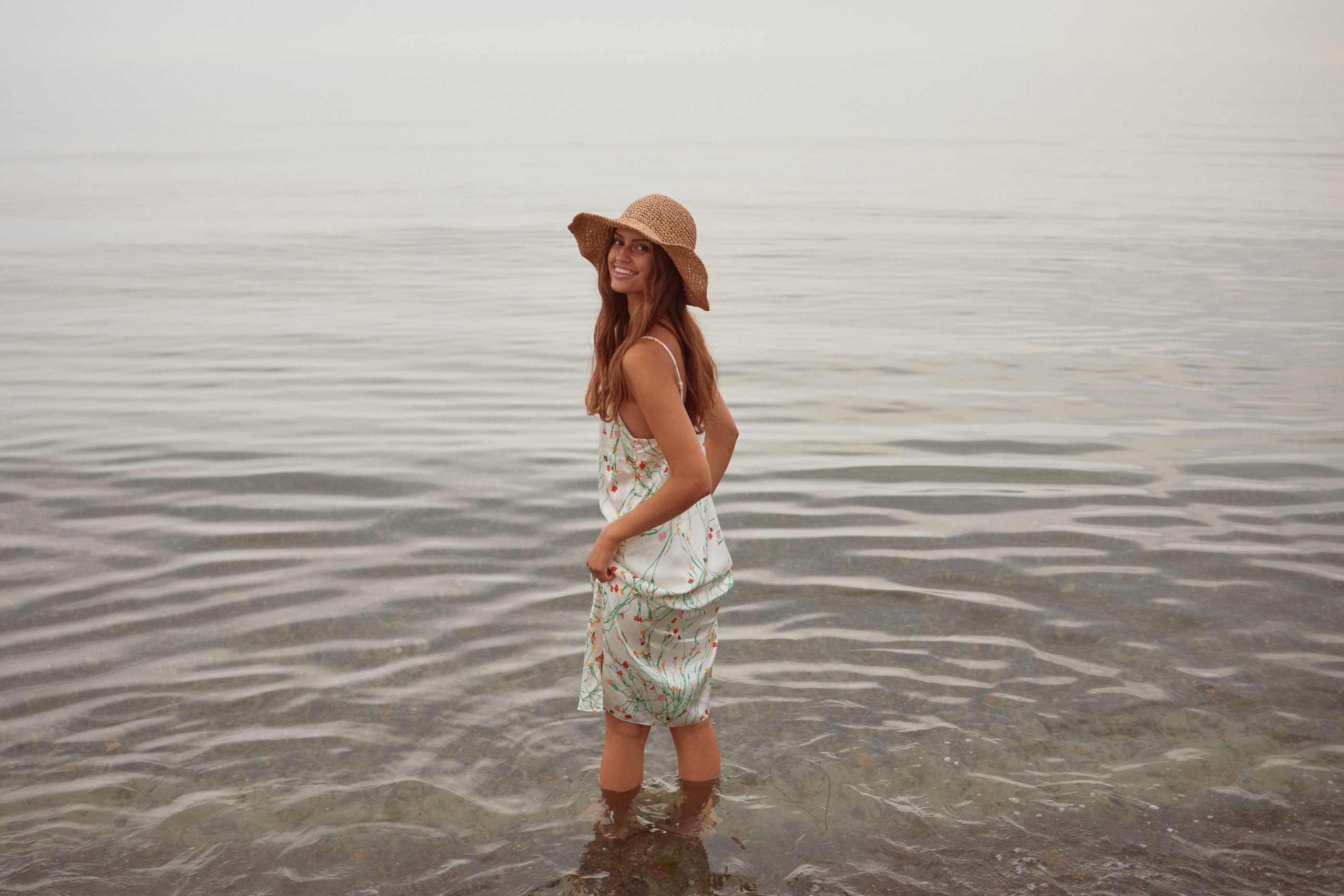Woman in floral dress and straw hat standing in calm water, smiling.