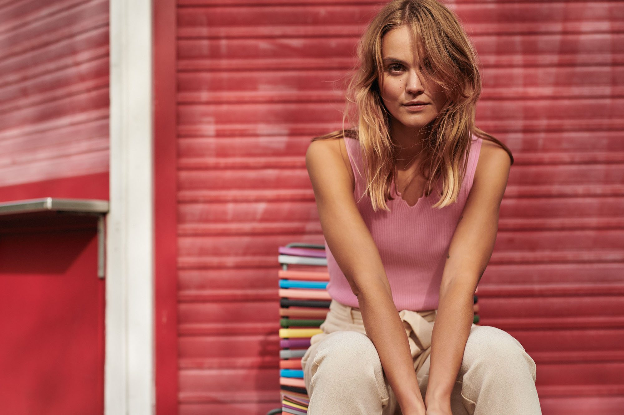 Blonde woman in pink top and beige pants sits on colorful bench against red wall.