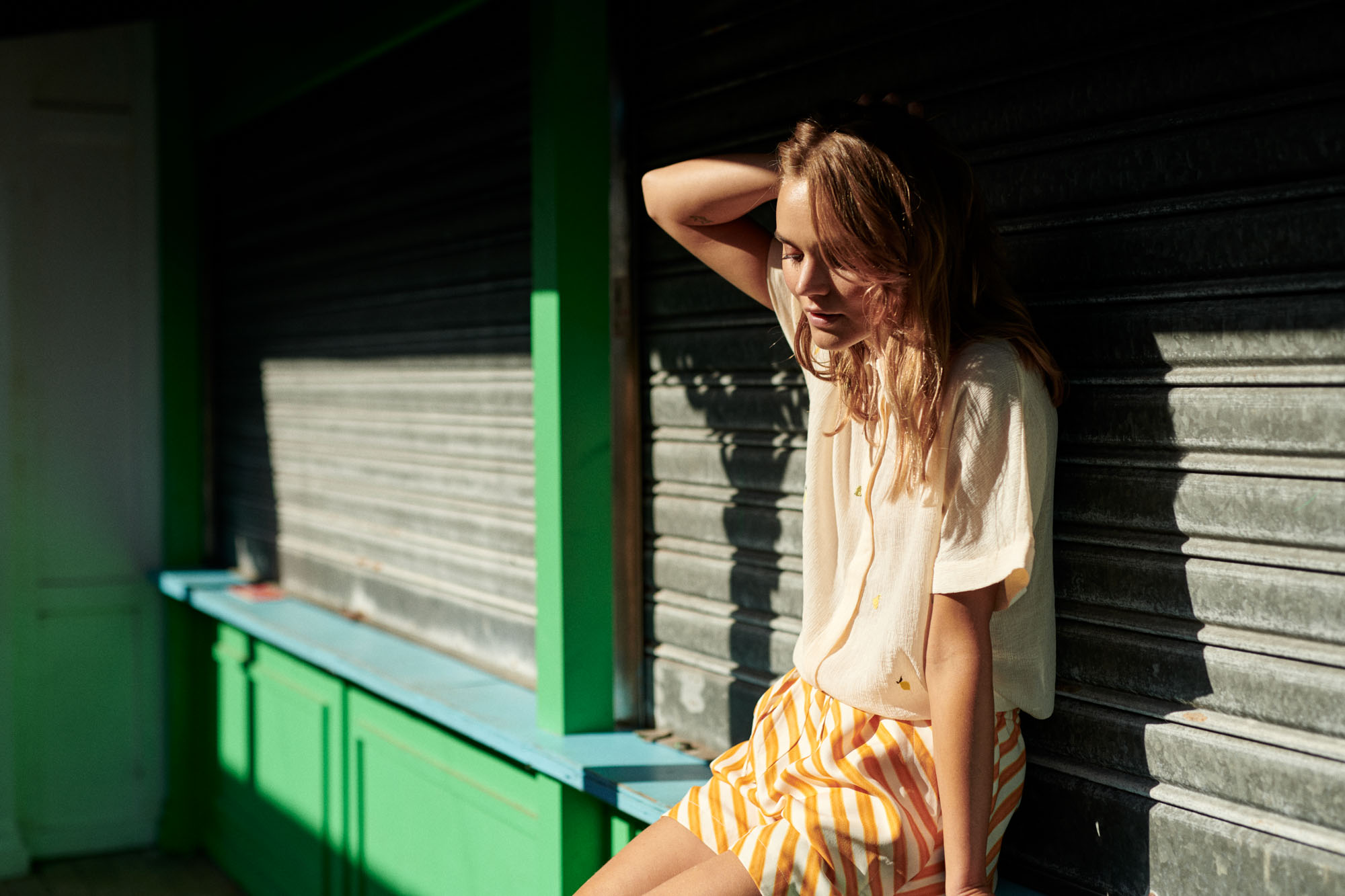 Woman in light top and striped shorts sits by dark corrugated wall in strong sunlight.