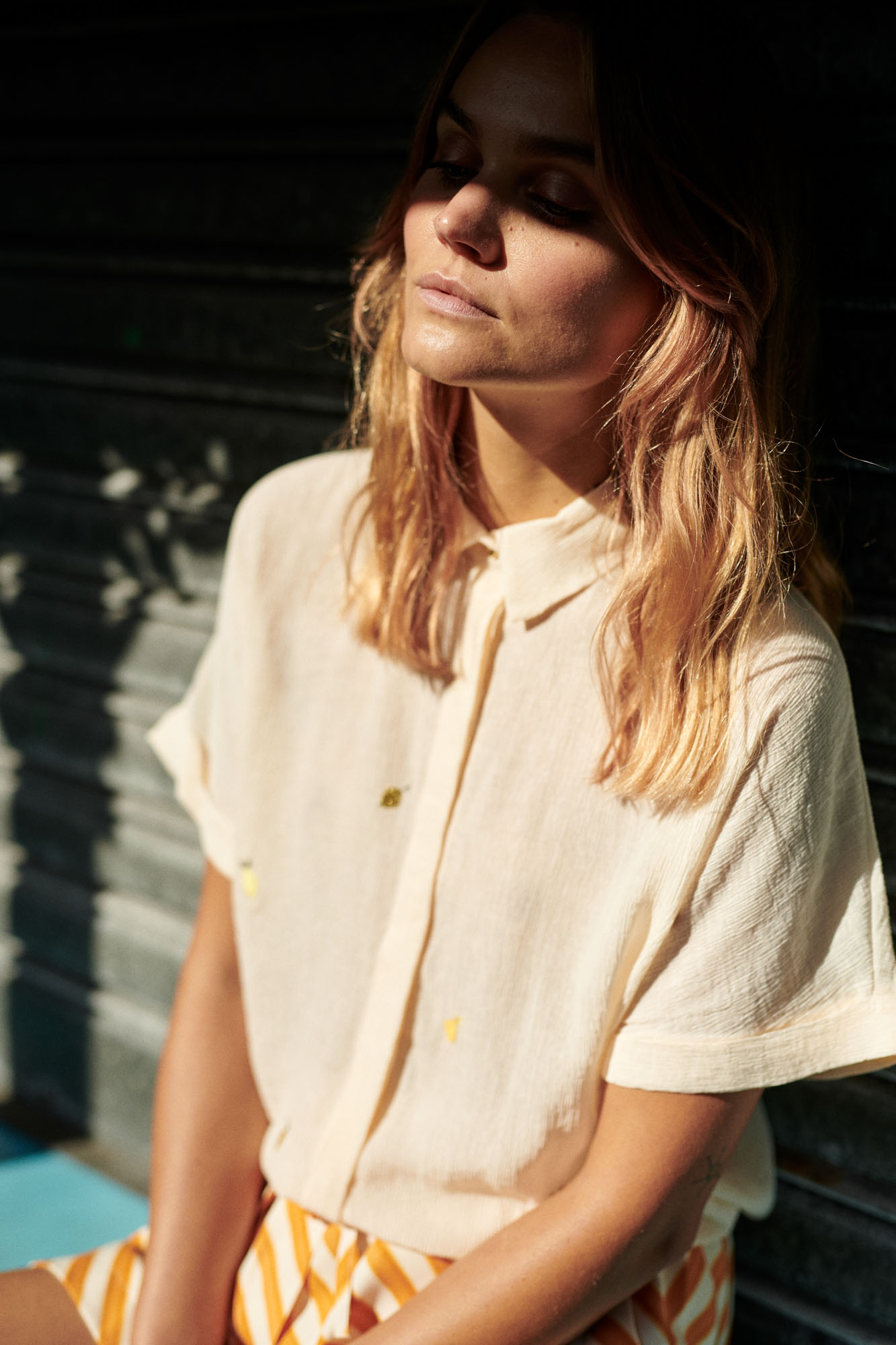 Relaxed woman in cream shirt and striped shorts, face illuminated by dappled sunlight.