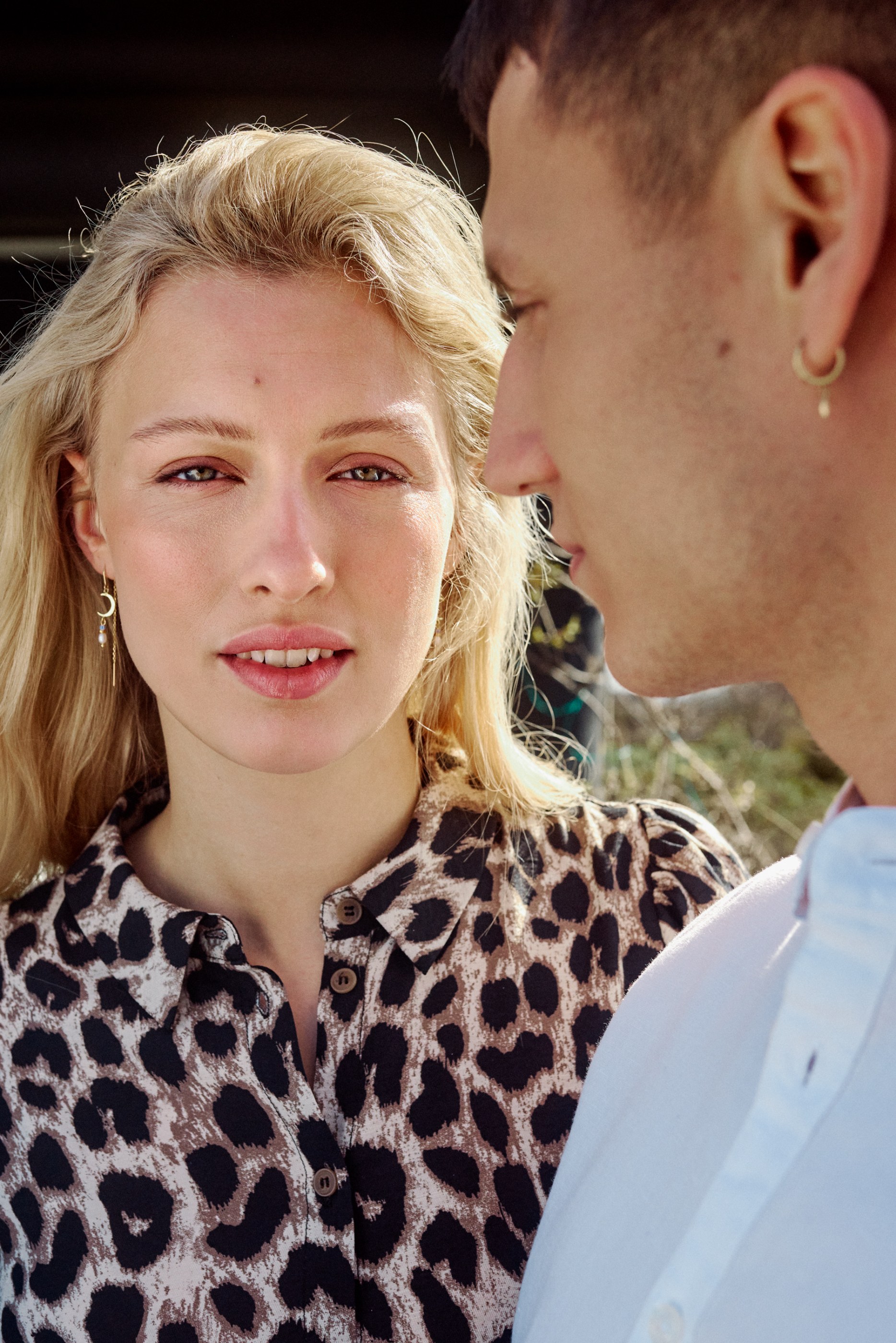 Blonde woman in leopard print shirt looks at viewer, man's blurred profile on right.
