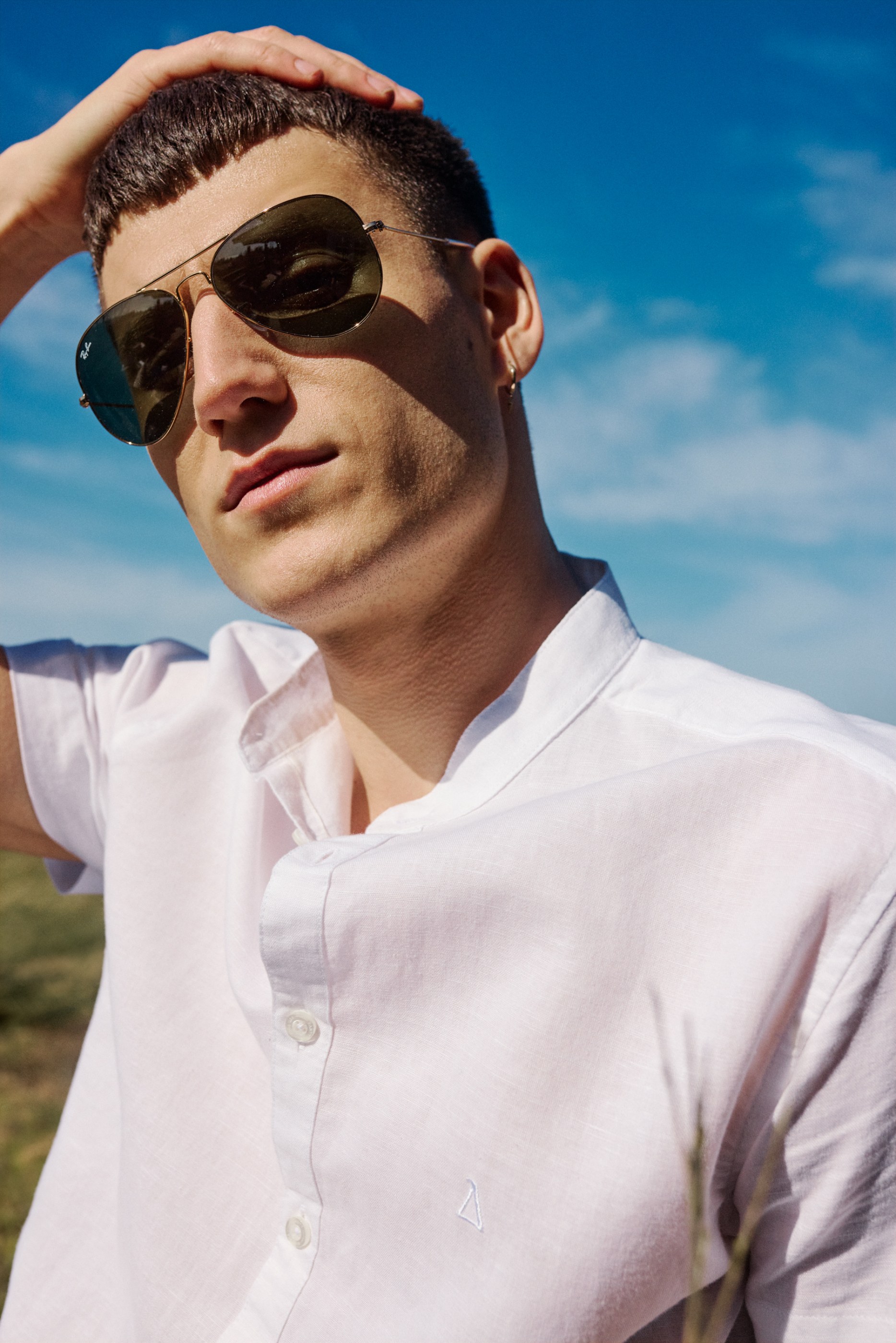 Young man in a white shirt and Ray-Ban sunglasses, with his hand in his hair against a blue sky.