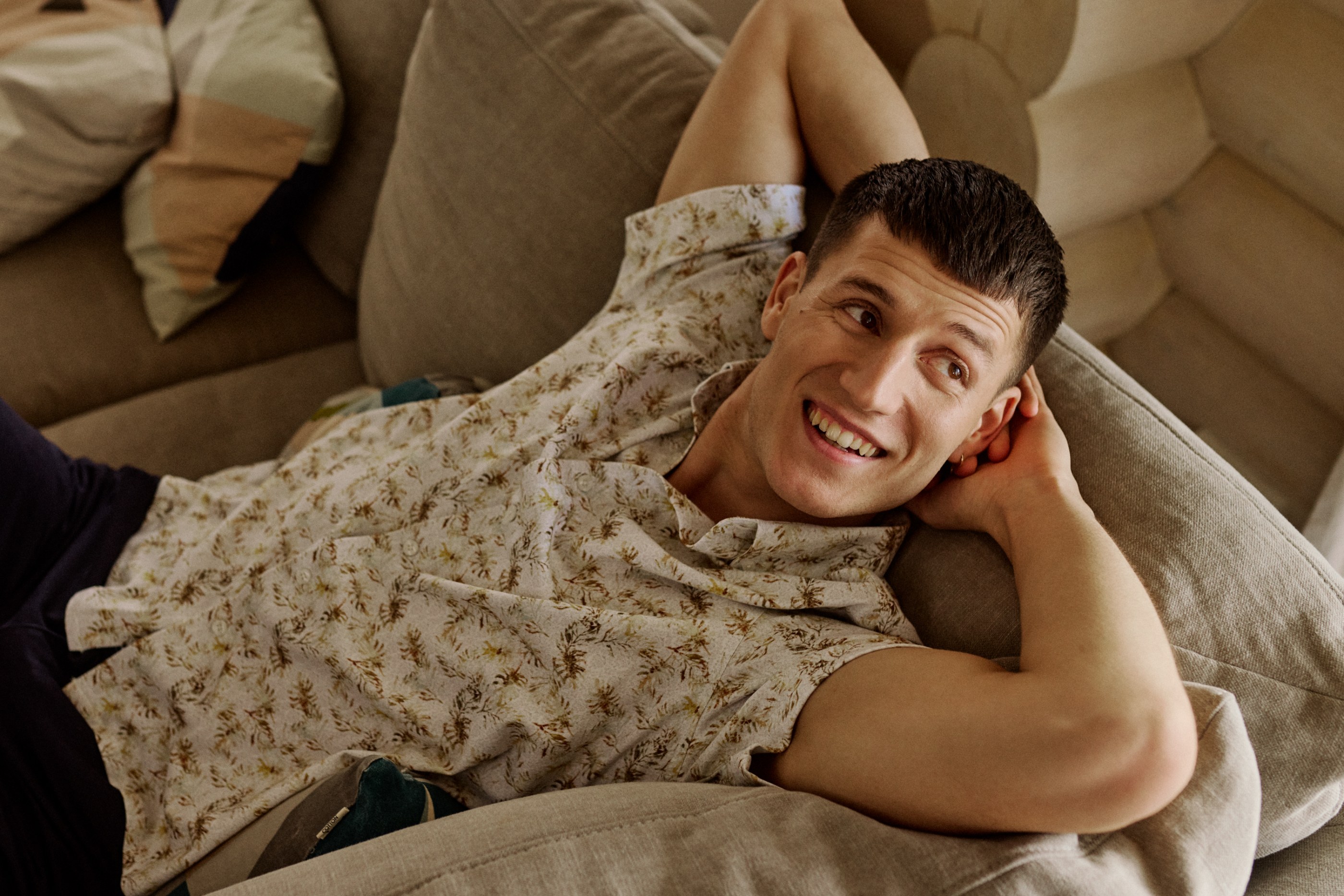A smiling man with a patterned shirt lies on a couch, looking to the side.