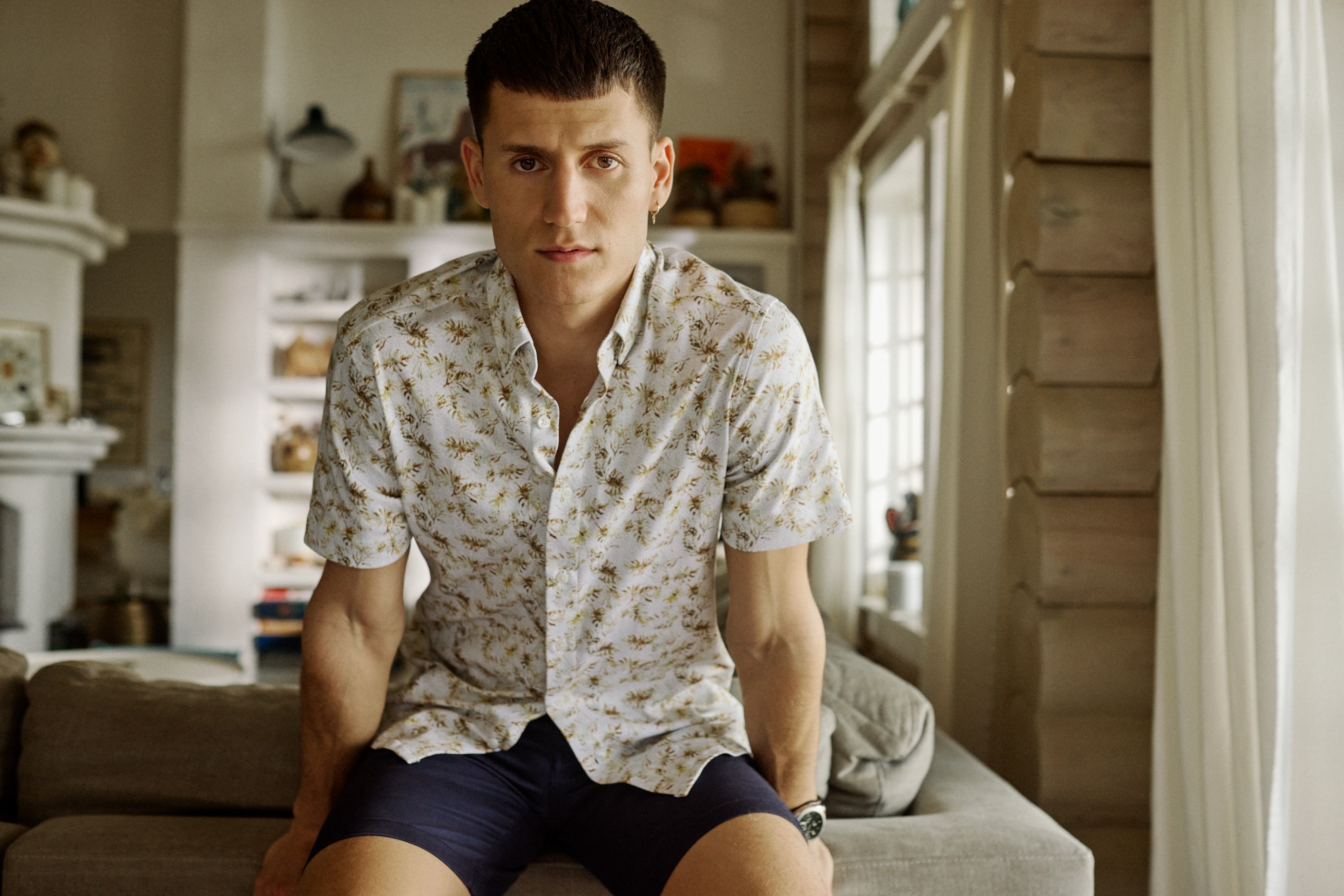 Young man in patterned shirt and navy shorts sits on a couch in a bright room.