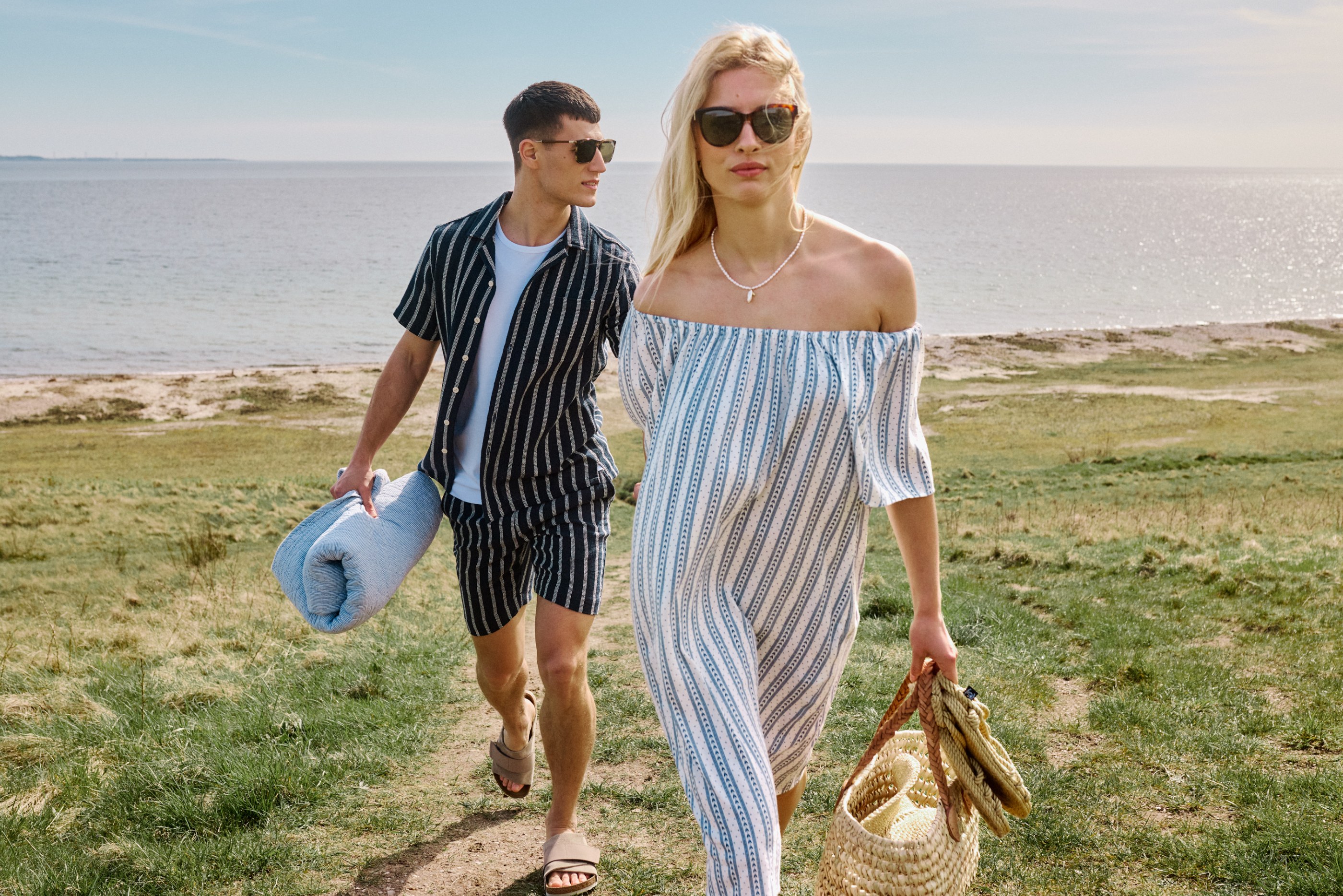 Couple in striped summer outfits walks a grassy path by the ocean on a sunny day.