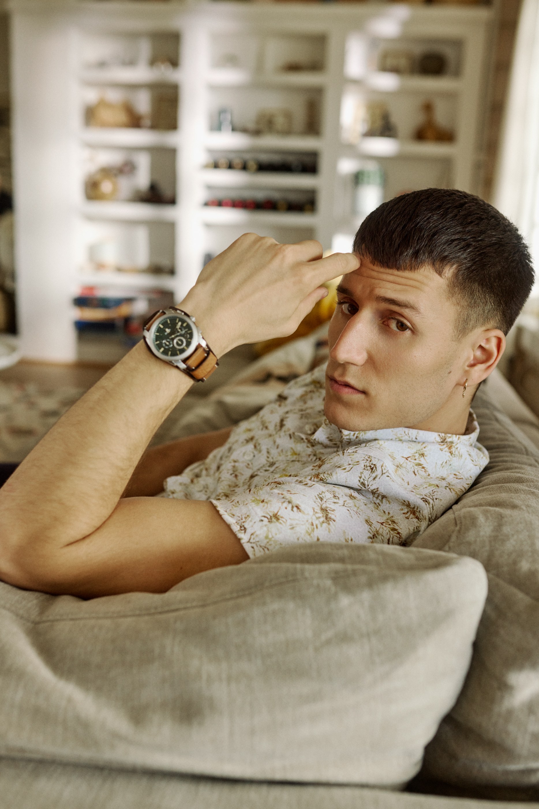 Young man in a patterned shirt and watch, lying on a couch, finger on forehead.