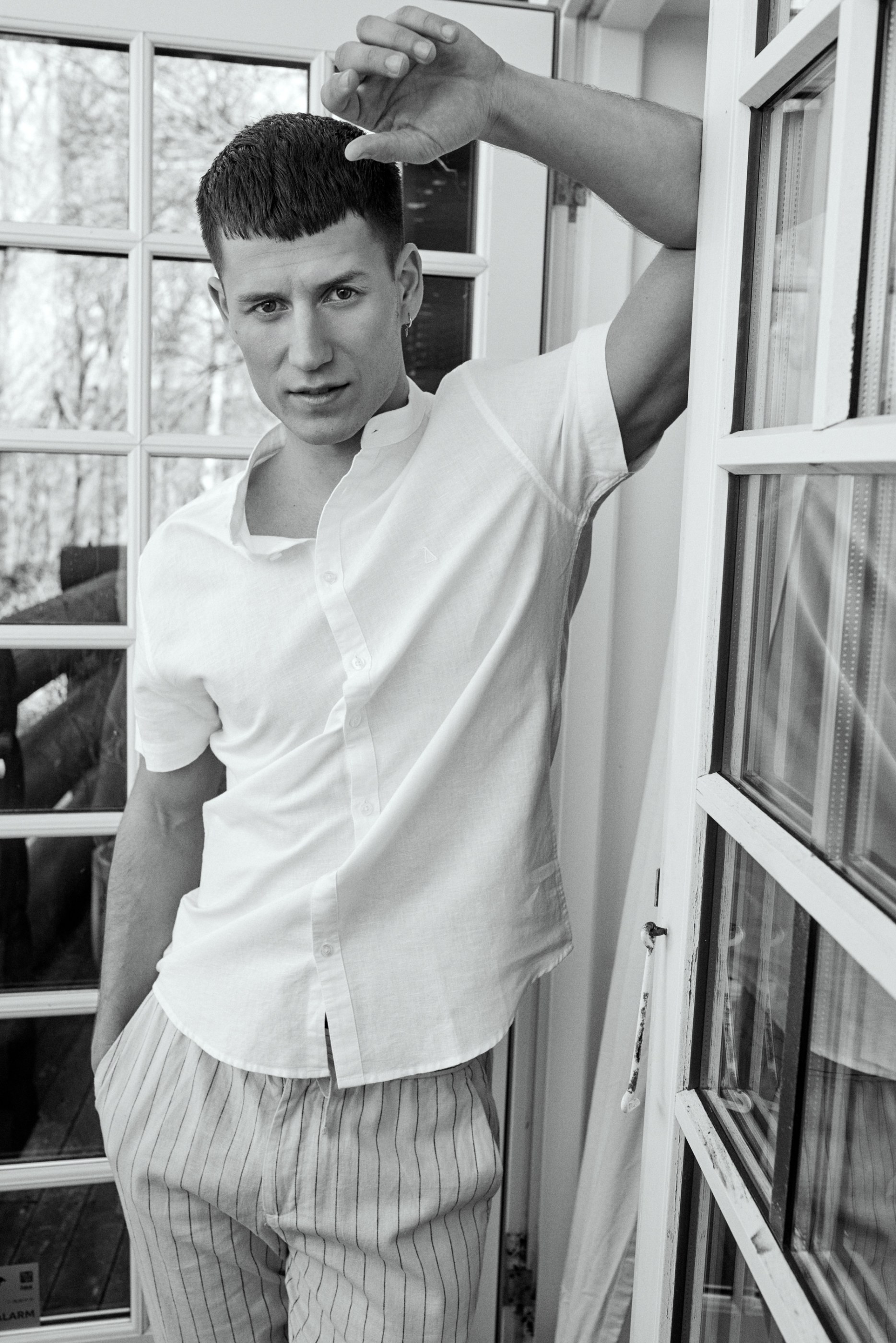 Young man in white shirt and striped pants leaning against a window frame, looking at camera.