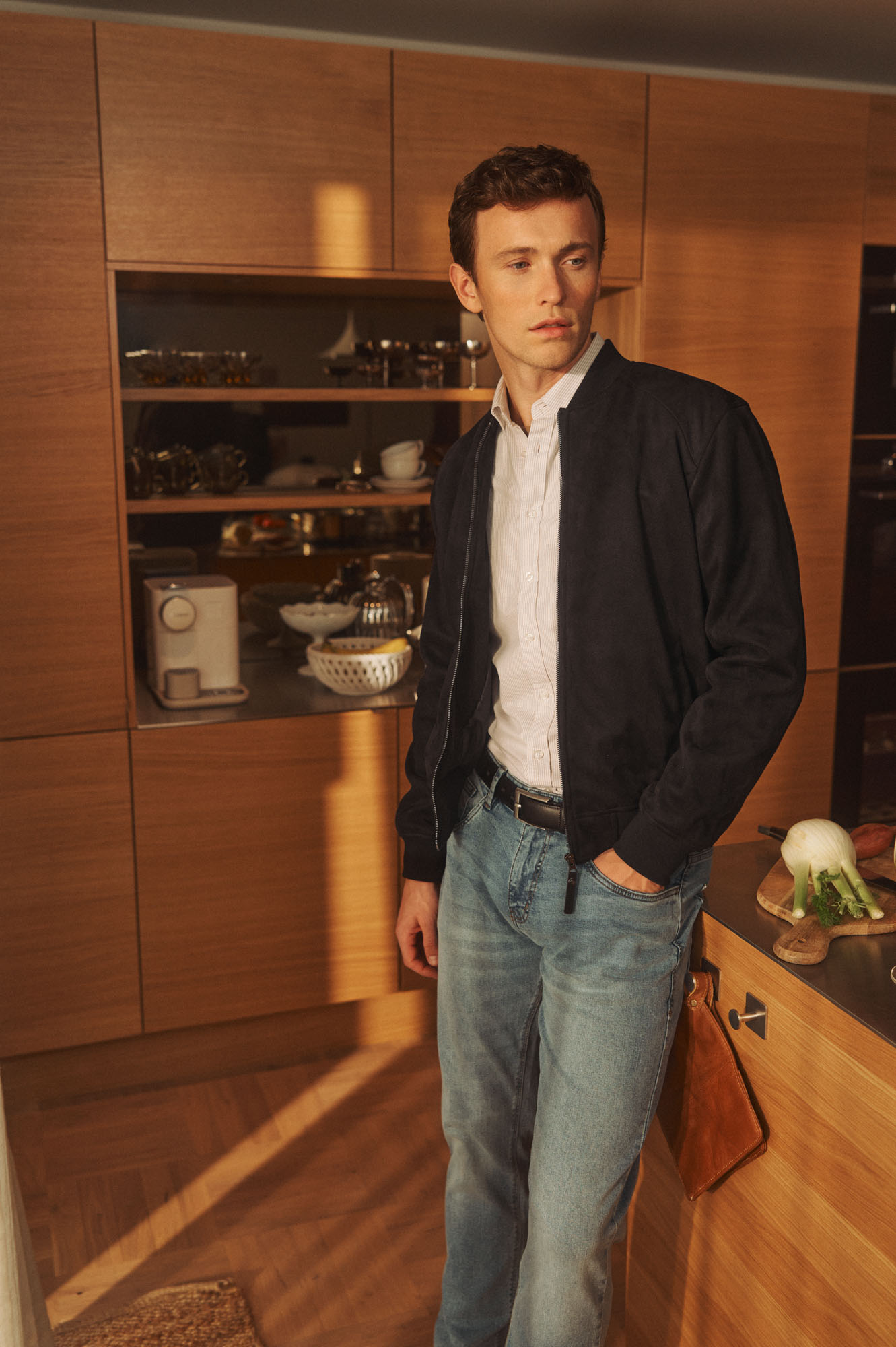 Young man in a navy bomber jacket and jeans standing in a warm-lit modern kitchen.