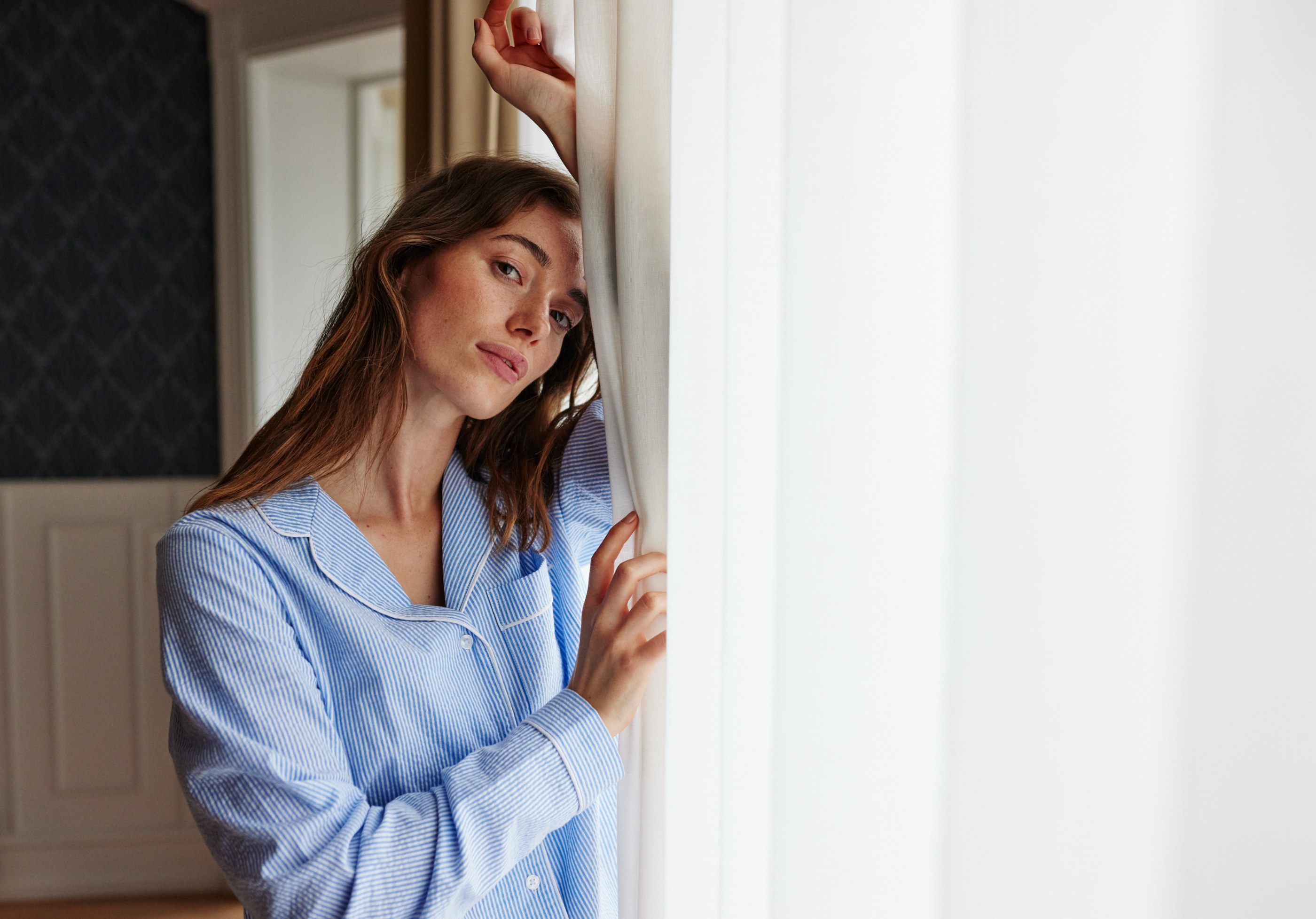 Woman in blue striped pajamas leaning against a sheer white curtain, looking calmly at the camera.