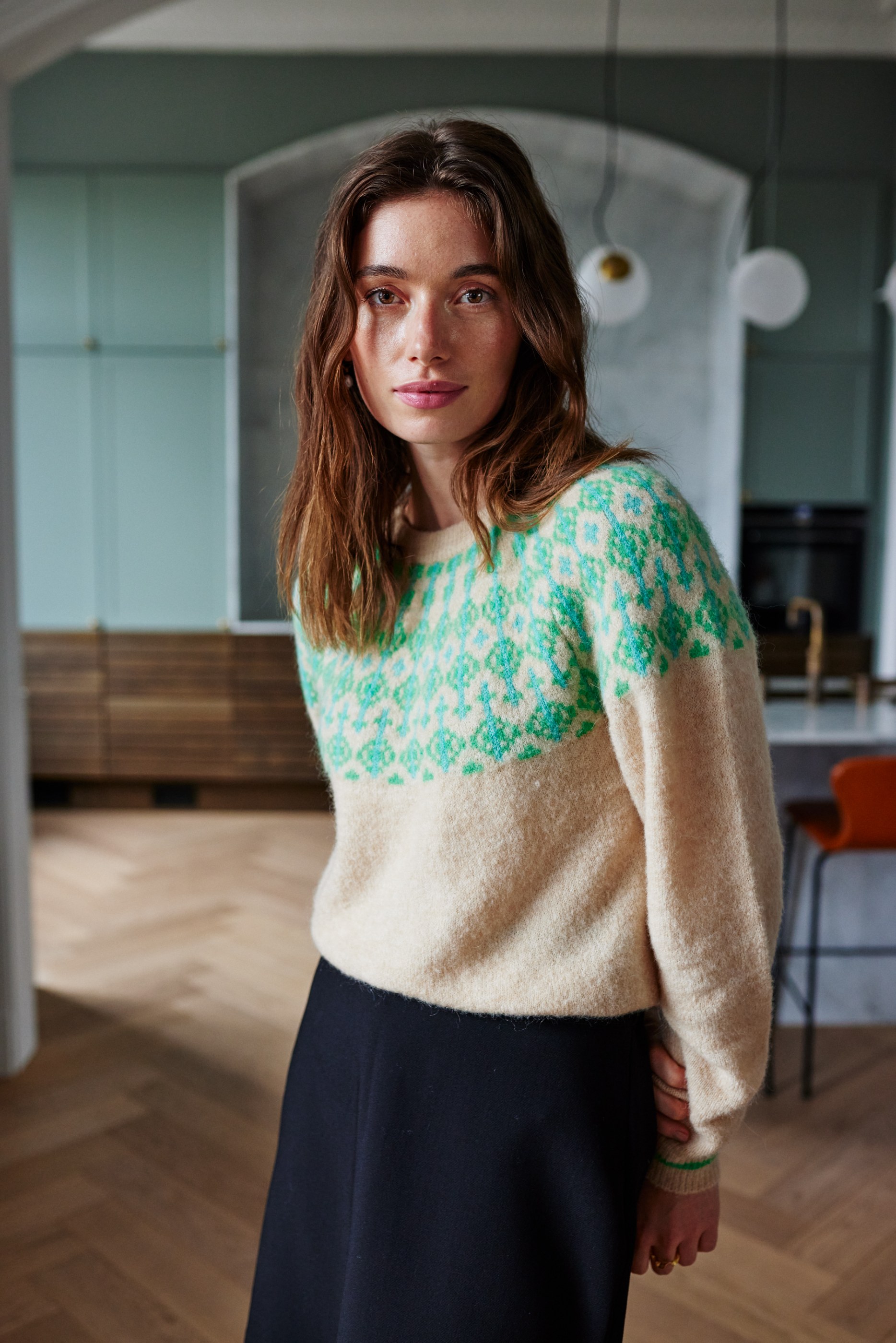 Brunette woman in a cream and green fair isle sweater and black skirt indoors.