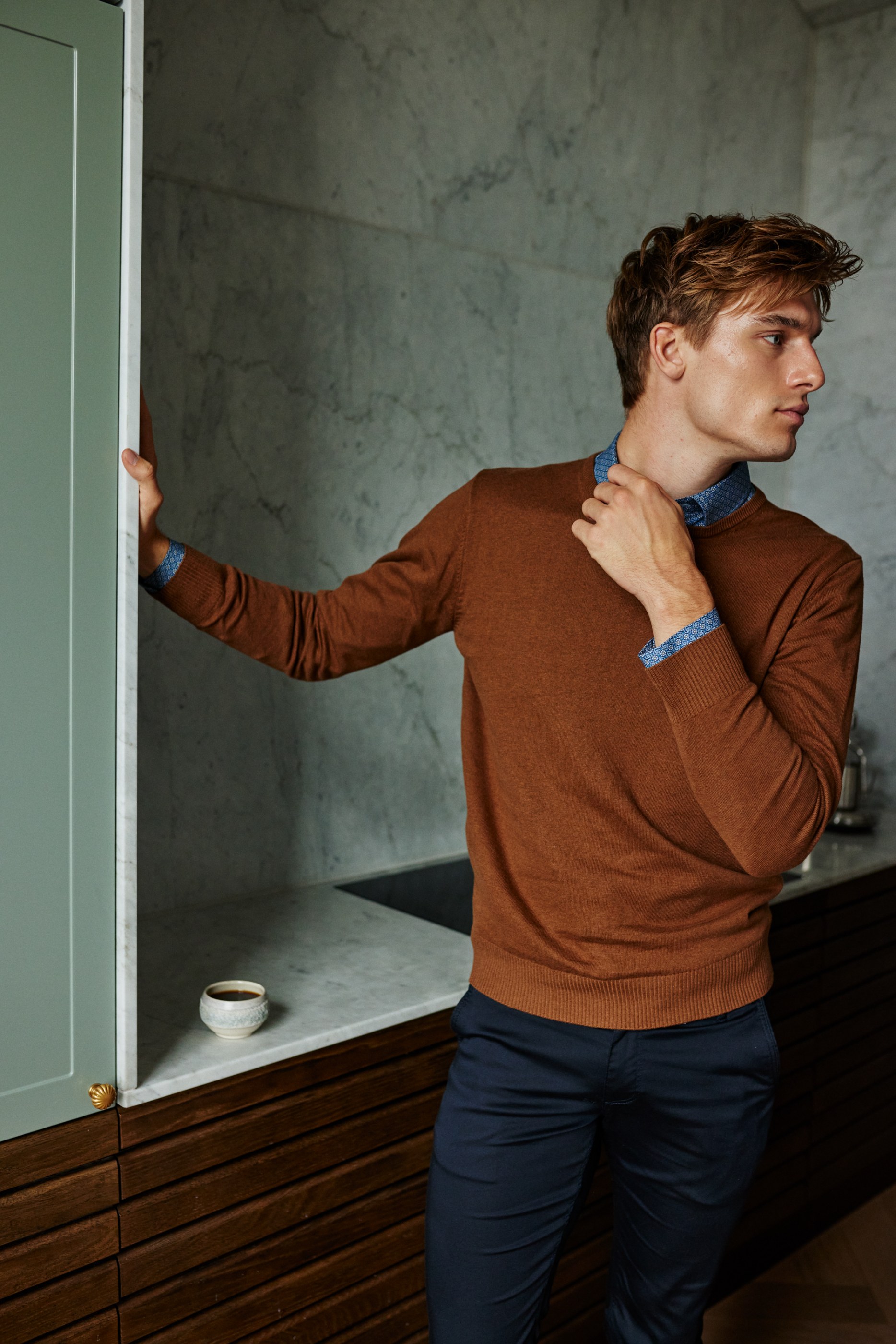 Man in a brown sweater and blue patterned shirt in a modern kitchen setting