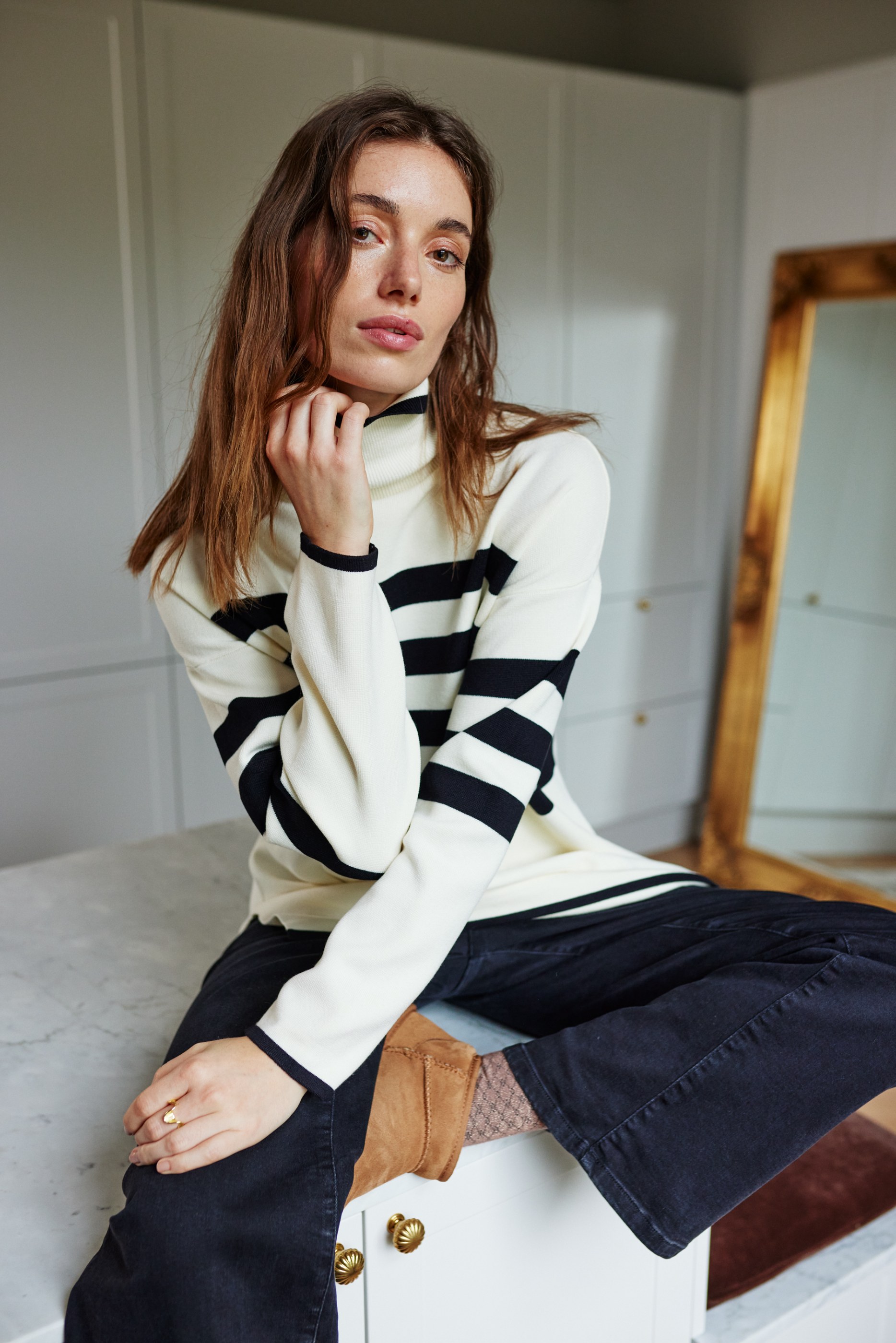 Woman in striped turtleneck sweater and dark pants sits on a white counter.