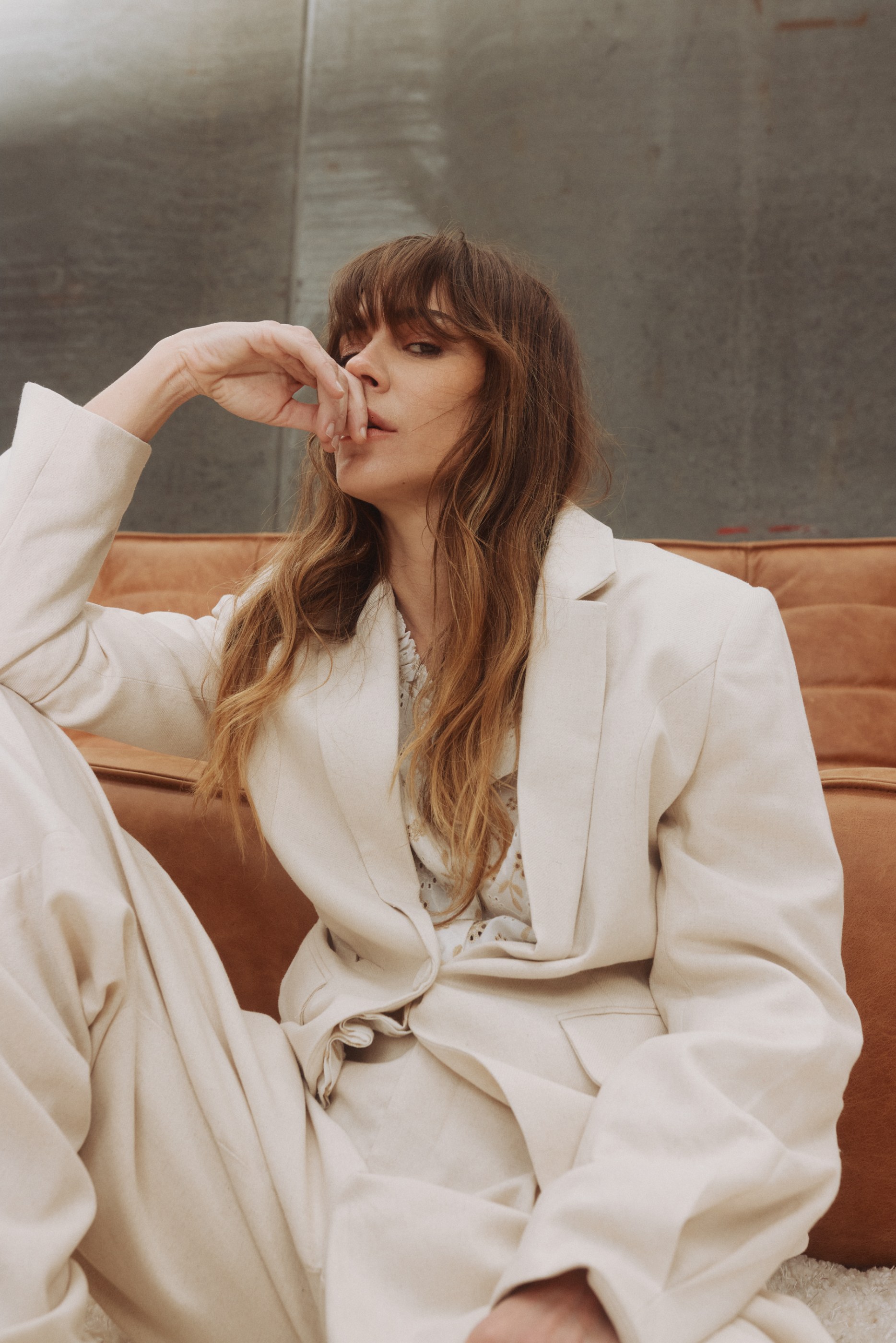 Woman with long brown hair and bangs wearing a cream suit, sitting pensive on a brown couch.
