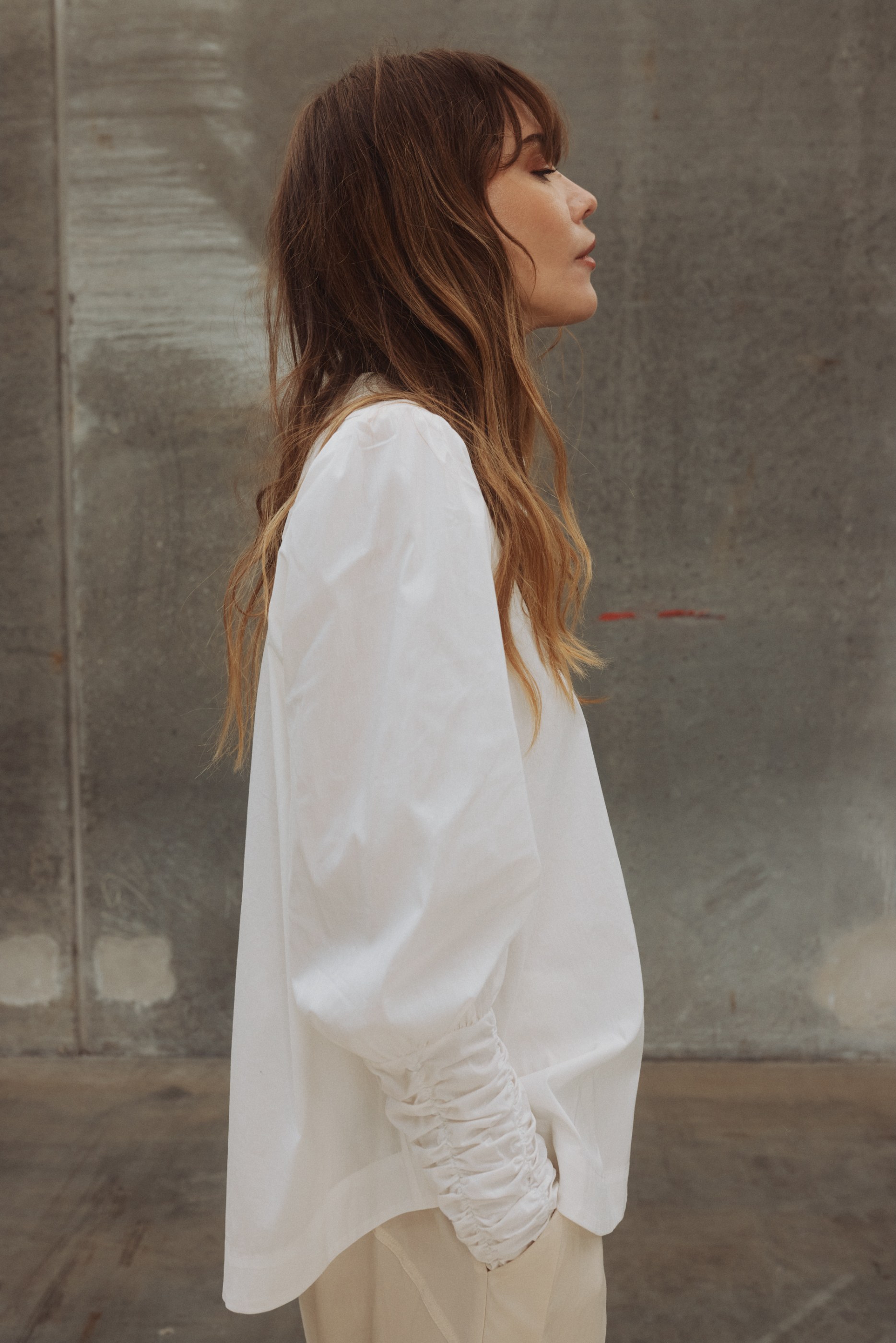 Profile of a woman with long brown hair, white ruched blouse, and beige pants.
