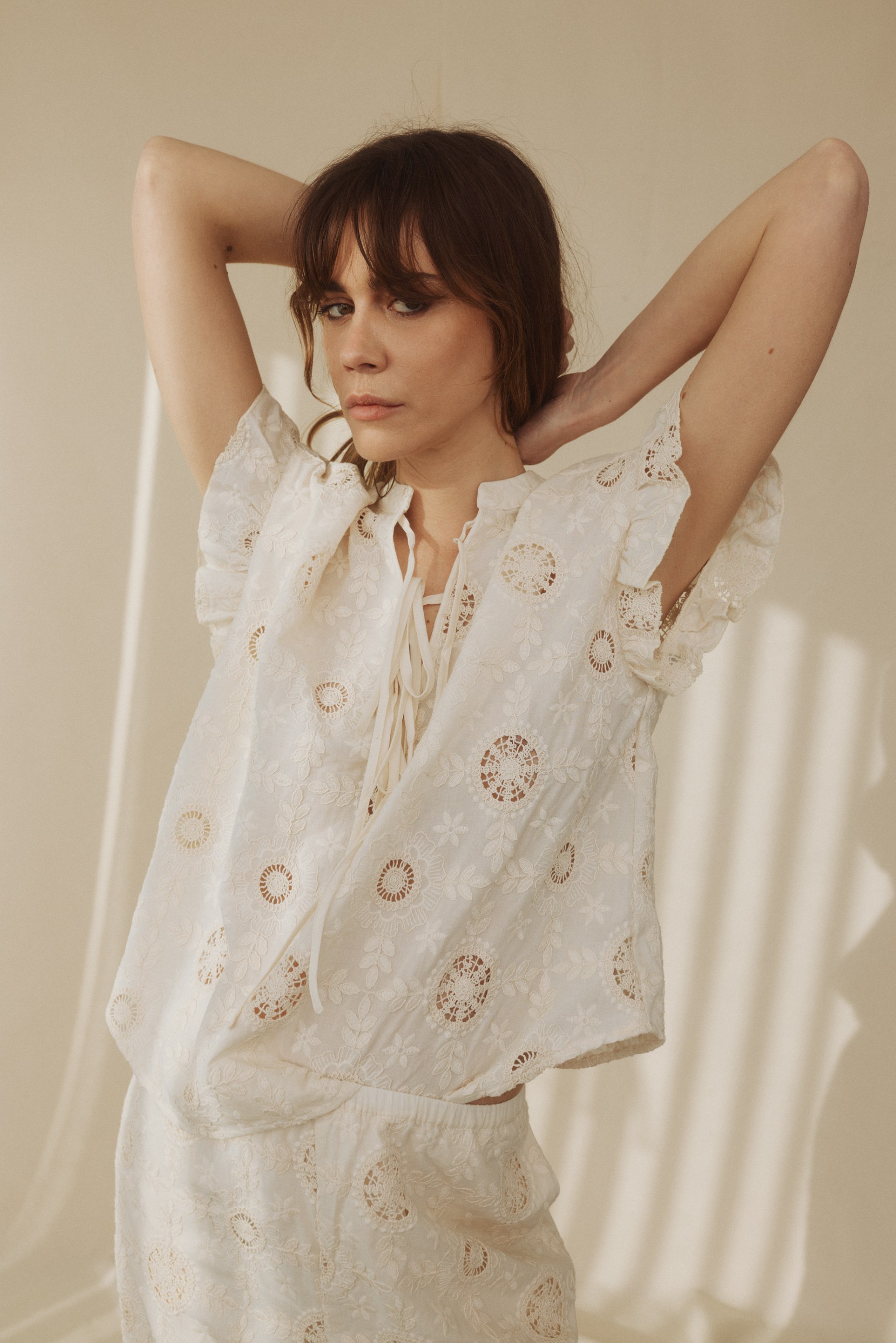 Woman with bangs in white embroidered top and shorts, hands behind head.