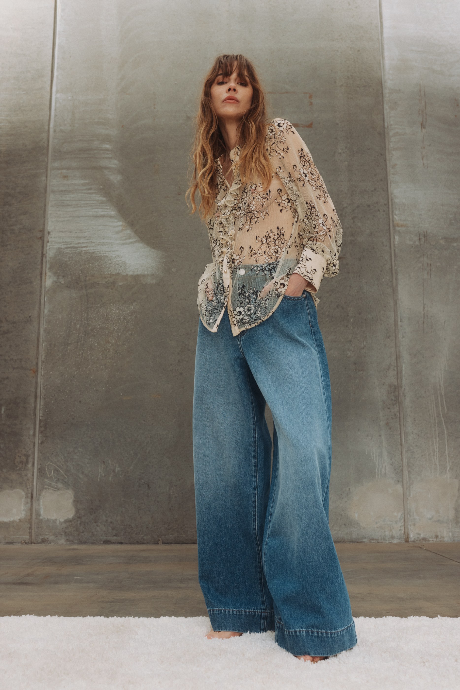Woman in sheer floral blouse and wide-leg jeans standing on a rug against a concrete wall.