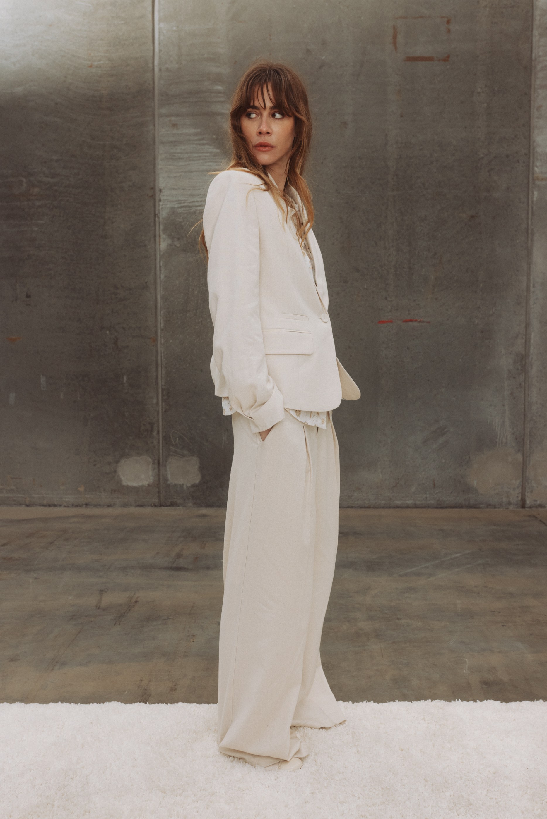 Woman in off-white blazer and pants looks over shoulder on a white rug against a grey wall.