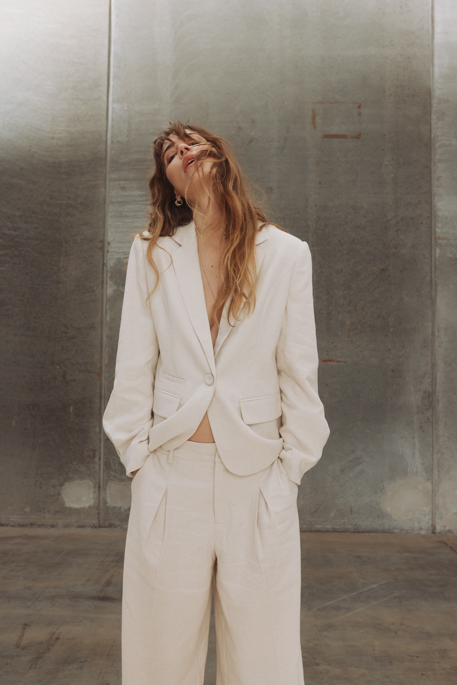 Woman in a light beige linen suit, hands in pockets, looking up against a concrete wall.