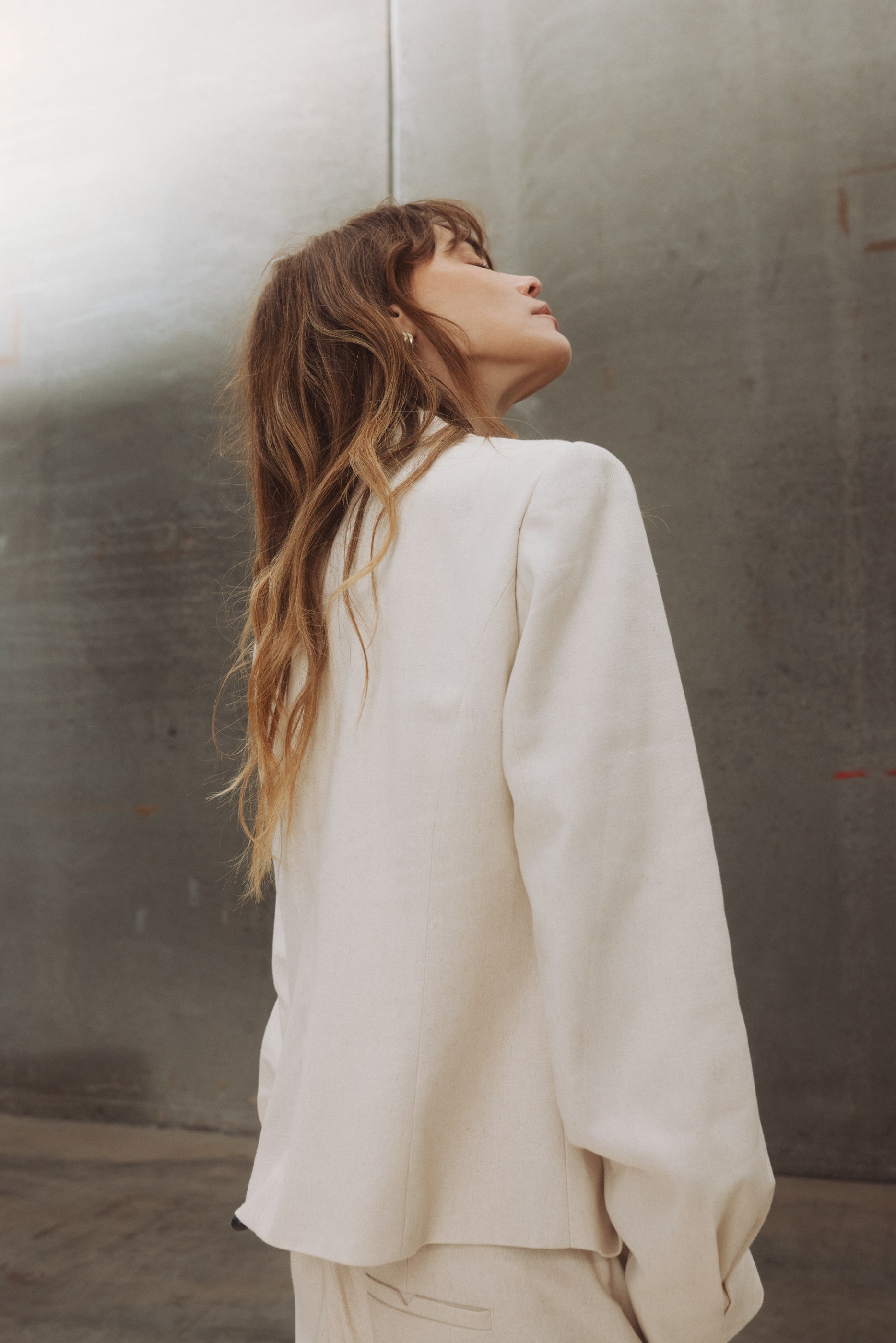 Woman with long wavy brown hair wearing a cream blazer looking upwards in profile.