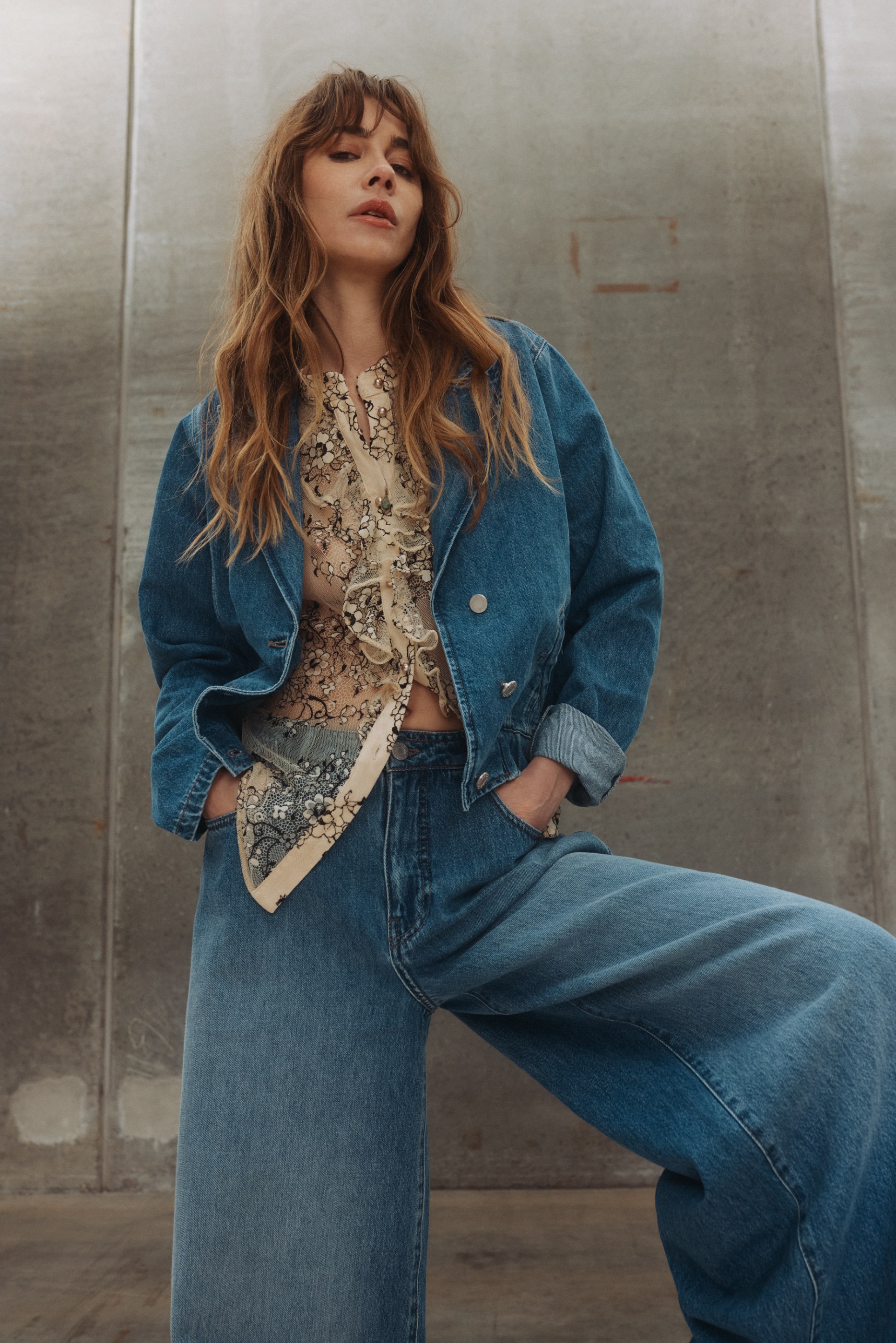 Confident woman with wavy hair in a denim jacket, ruffled lace top, and wide-leg jeans by a concrete wall.