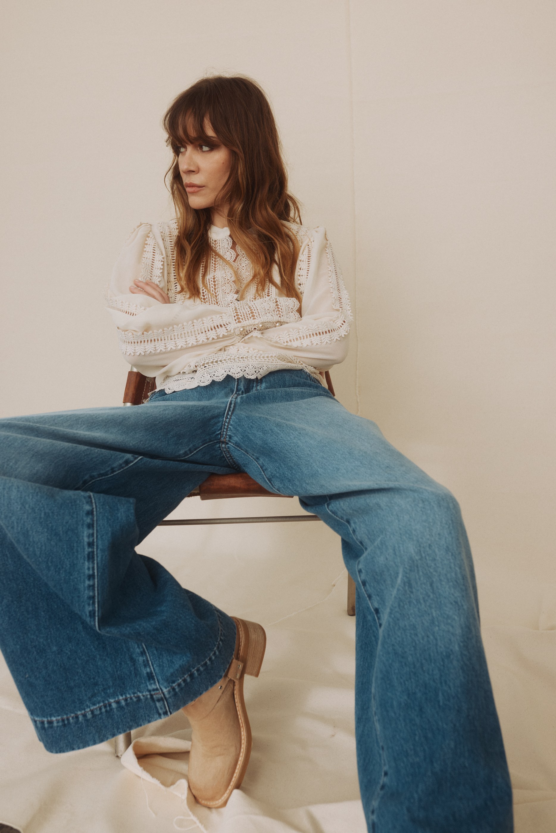 Woman in white lace top and wide blue jeans sits looking left, arms crossed.