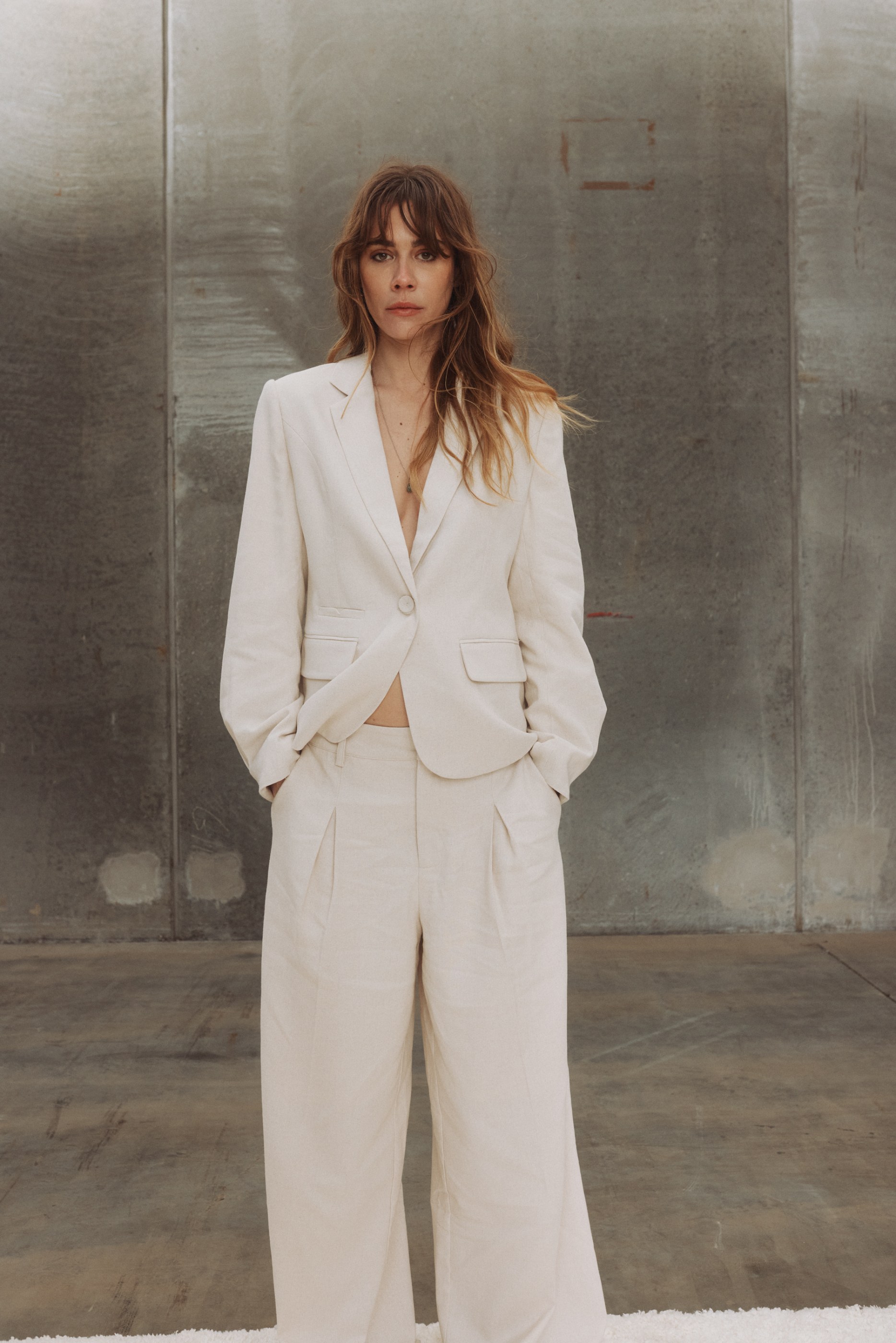 Woman in a cream linen suit, hands in pockets, looking at camera against a grey wall.