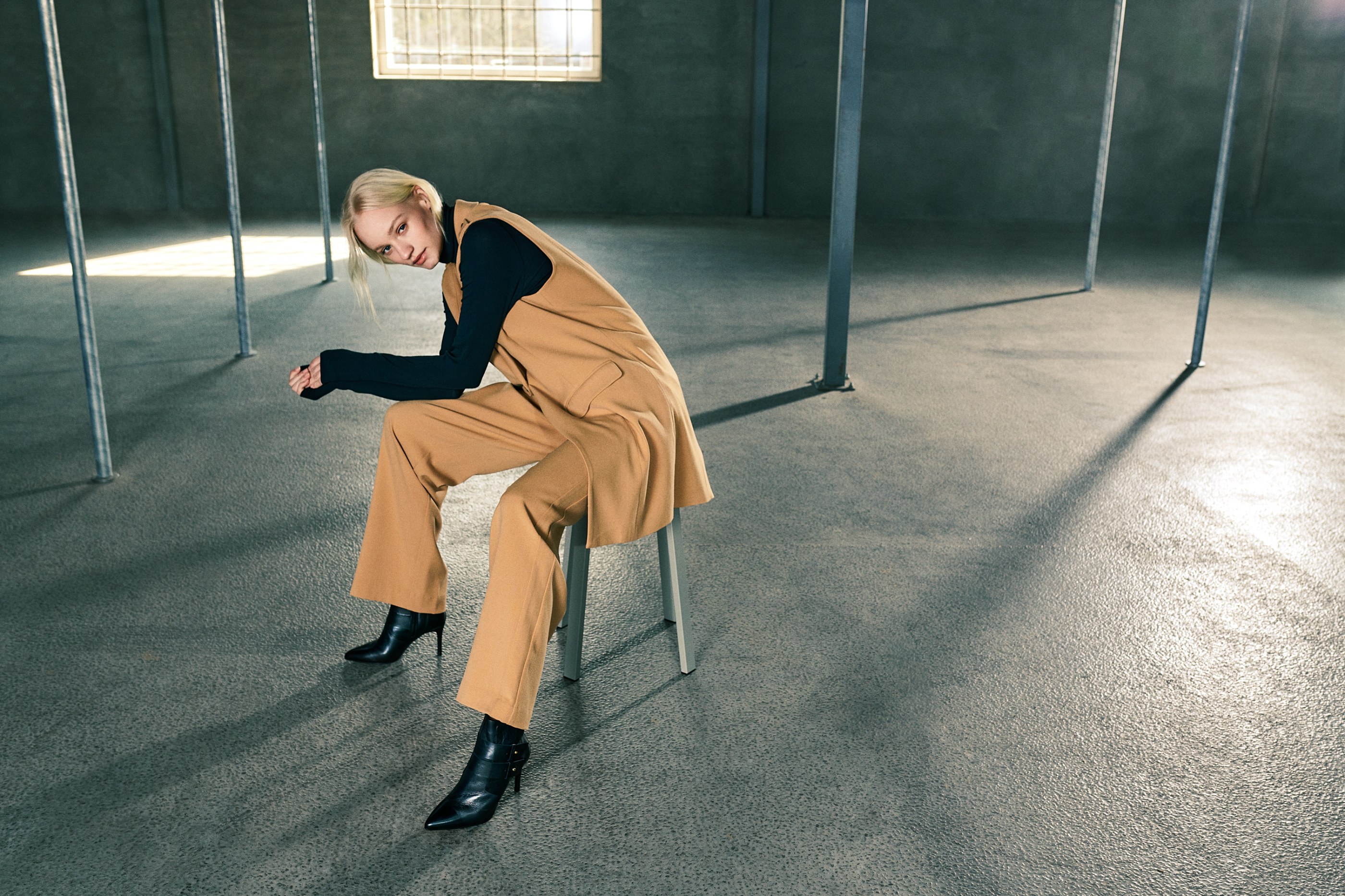 Blonde woman in black turtleneck, camel pantsuit, on stool in industrial concrete room.