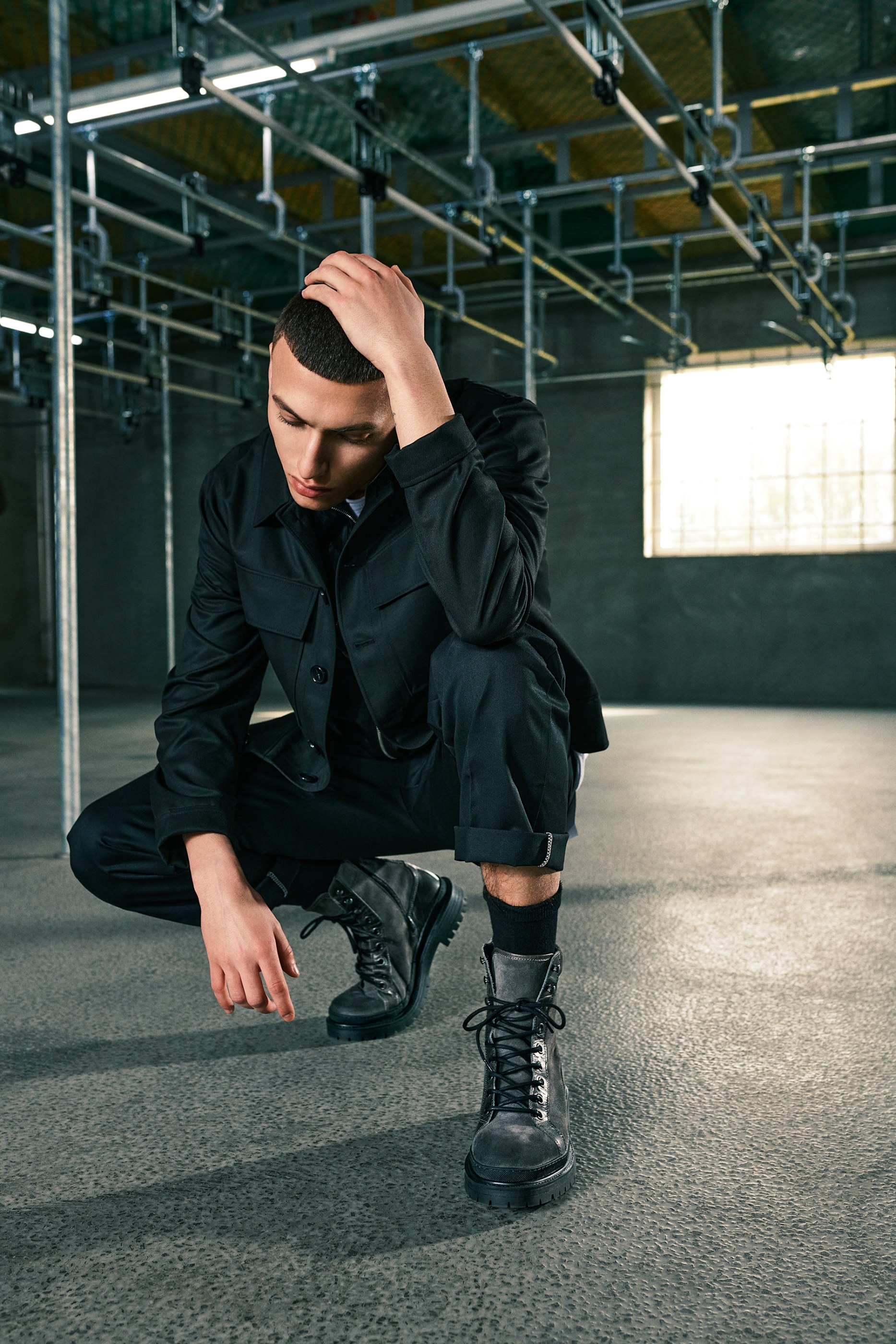 Man in dark clothes and boots crouches, hand on head, in industrial setting.