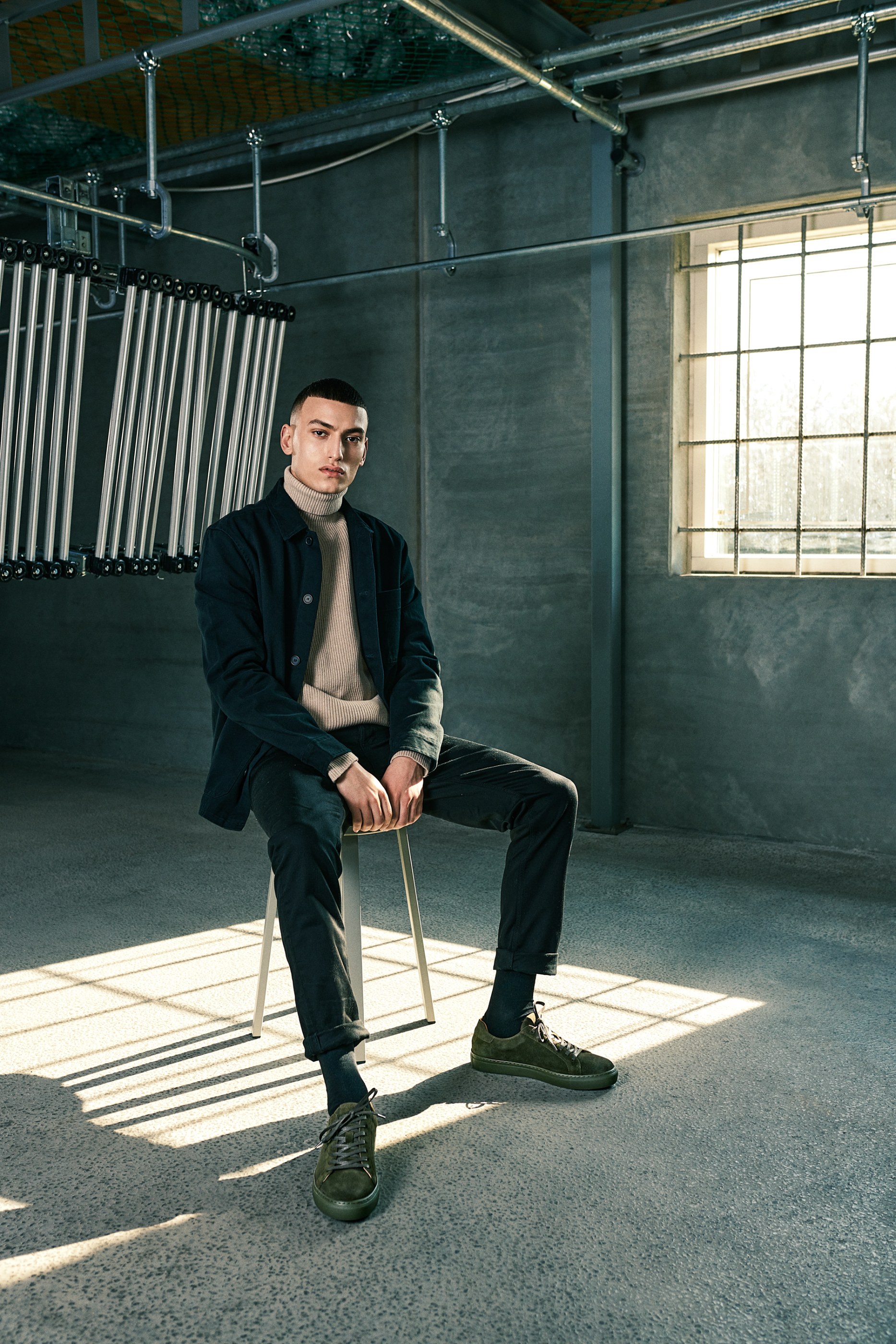 Man in turtleneck and jacket sits on stool in industrial room, sunlight casting shadows.