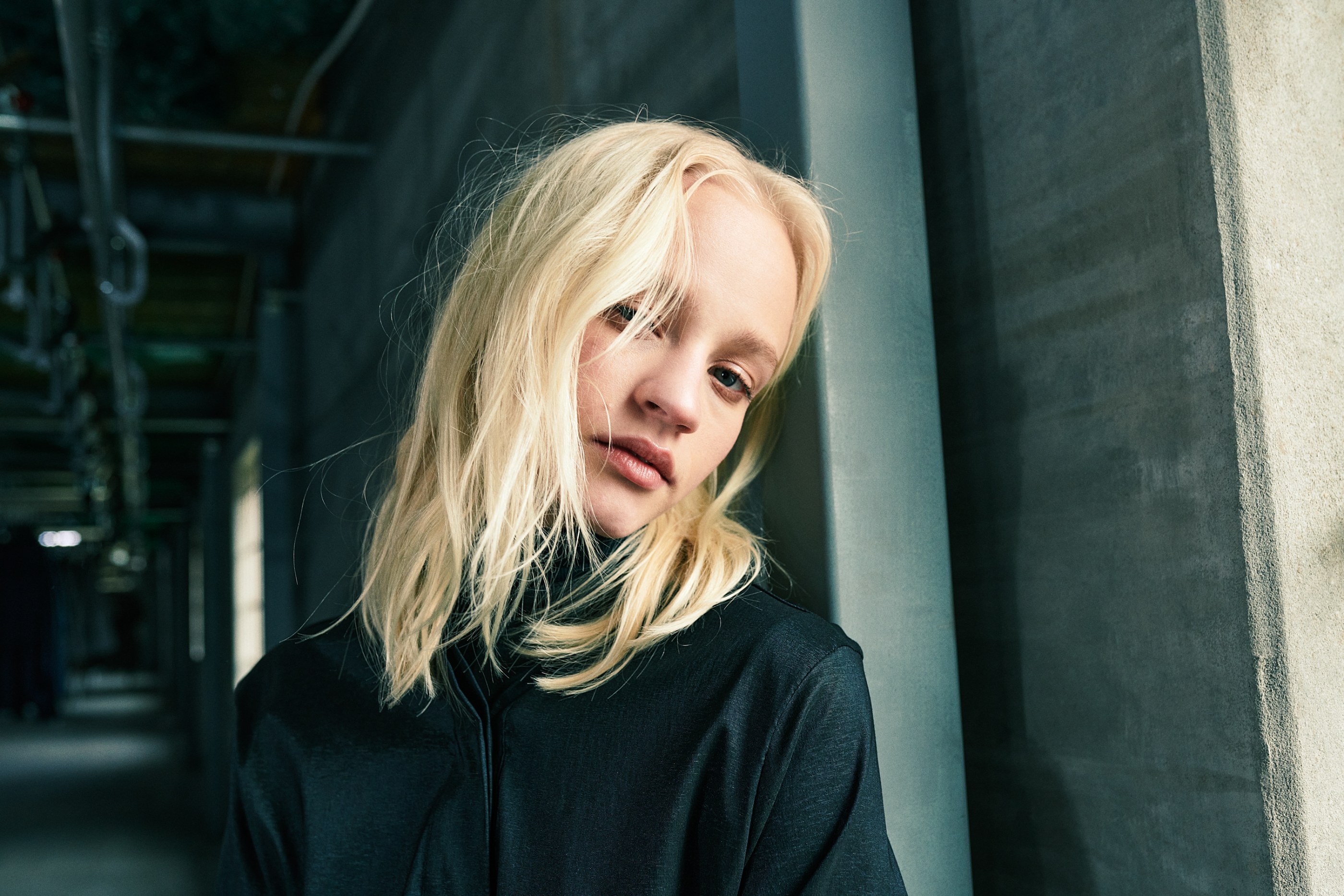 Contemplative blonde woman in black shirt leaning against a sunlit concrete wall.
