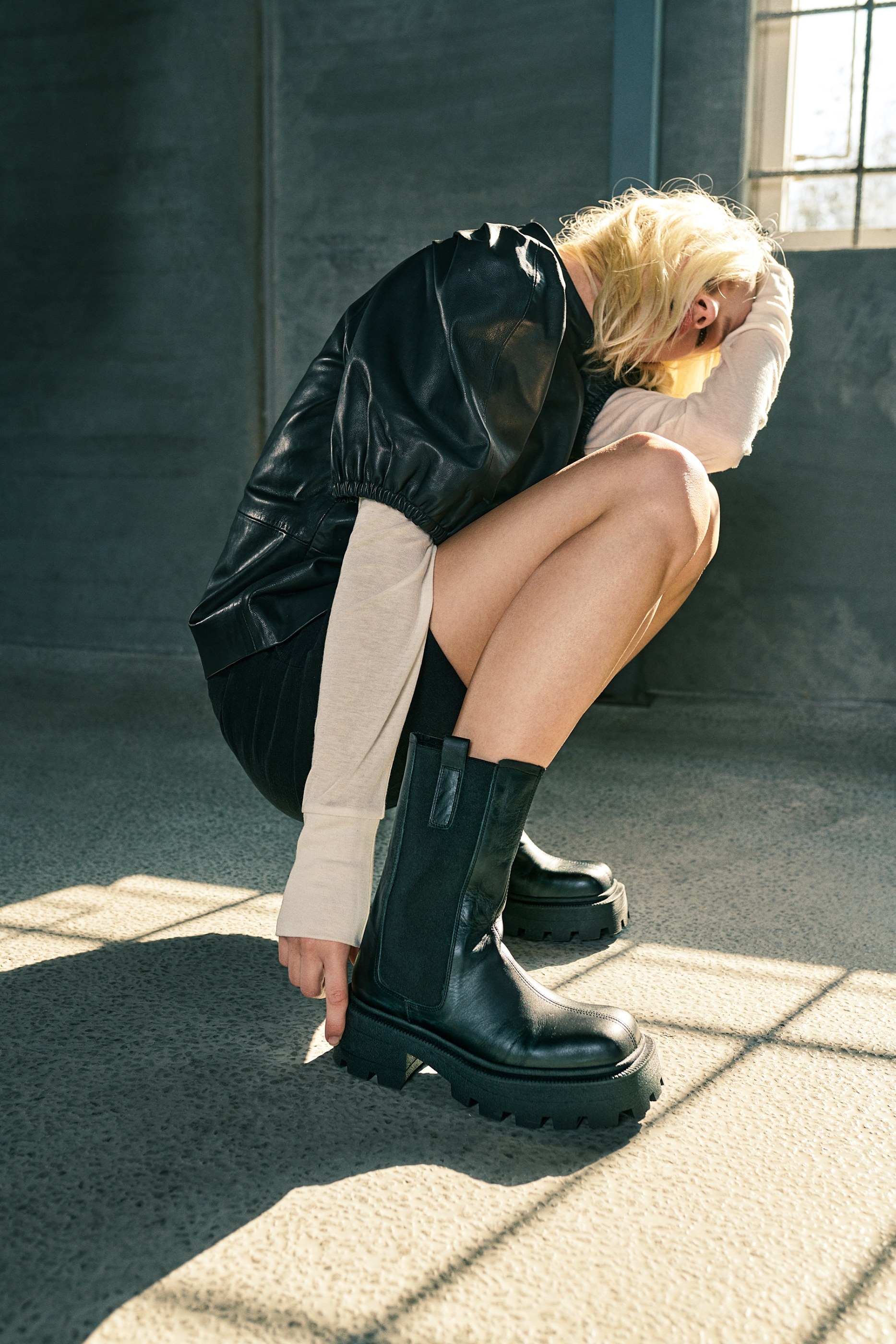 Person in black leather jacket and platform boots crouches on concrete floor with strong sunlight.