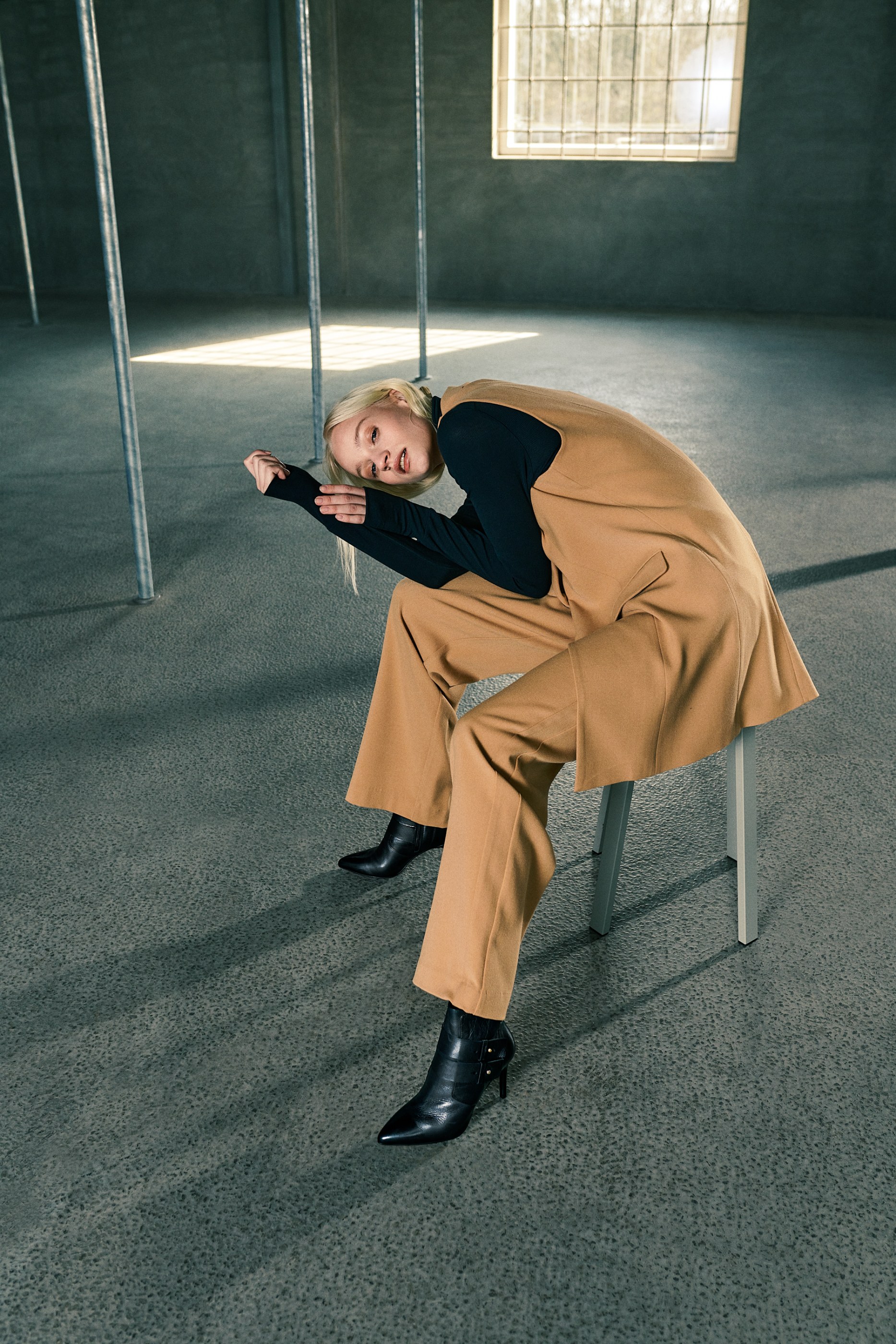 Blonde person in tan outfit and black top leaning in an industrial minimalist room.