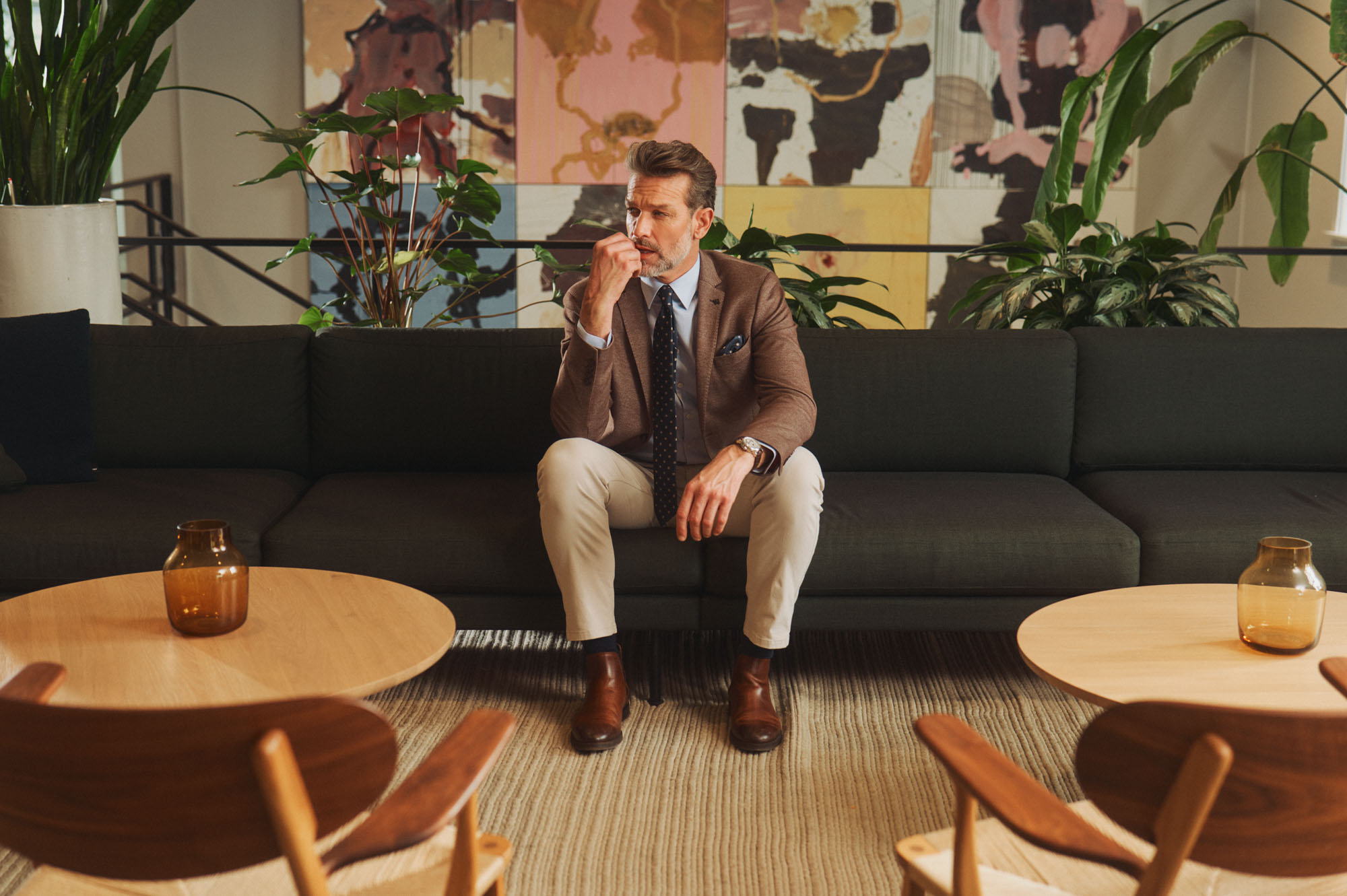 Thoughtful man in a brown blazer sitting on a sofa in a stylish lounge with plants and artwork.