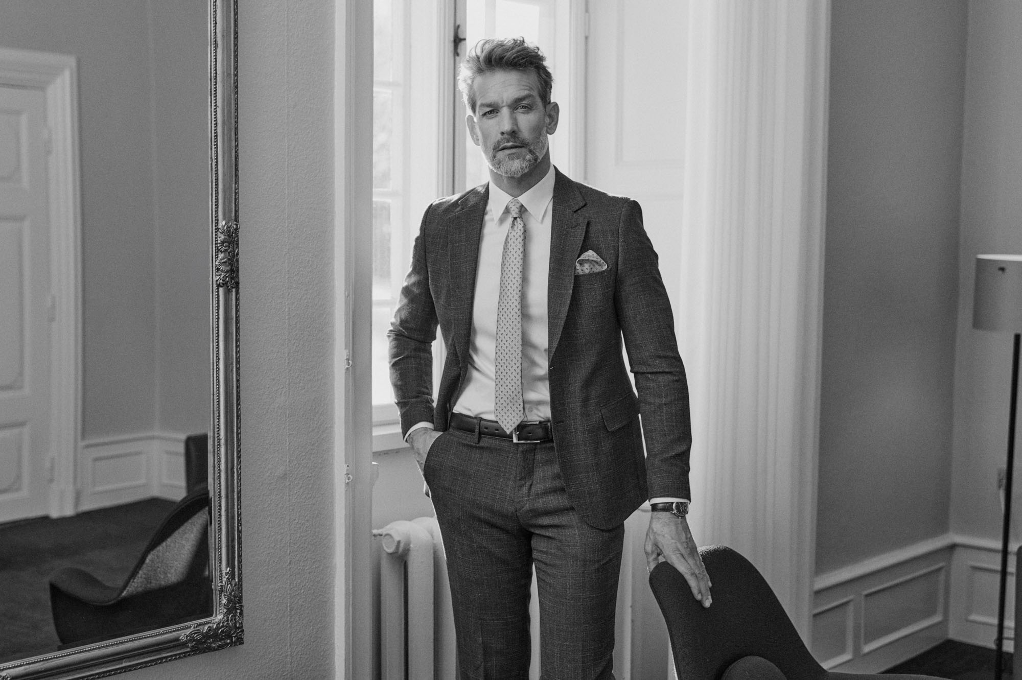 Black and white portrait of a stylish man in a tailored suit by a window and mirror.