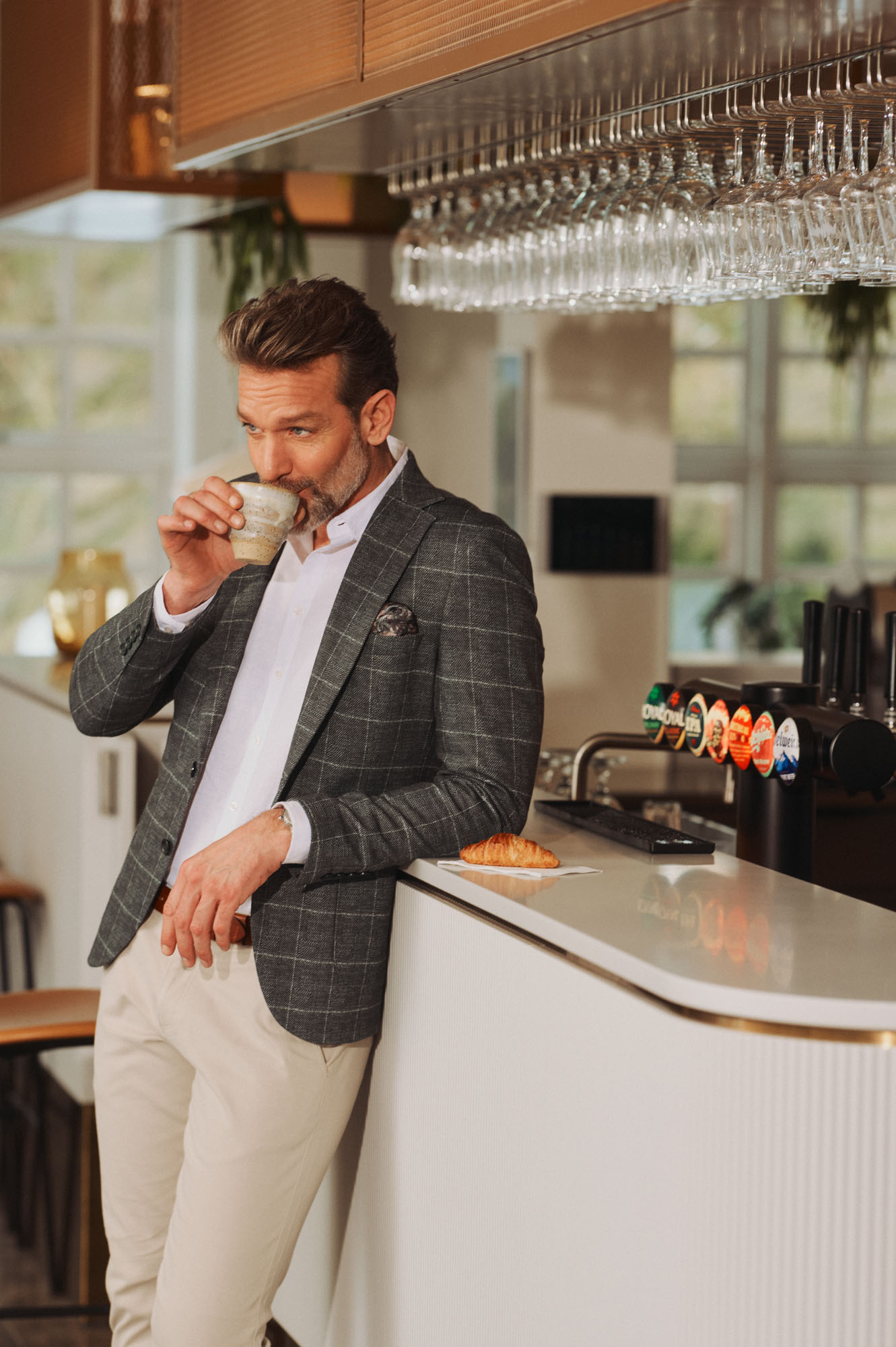 Man in a plaid blazer and white shirt sips coffee, leaning on a modern bar.