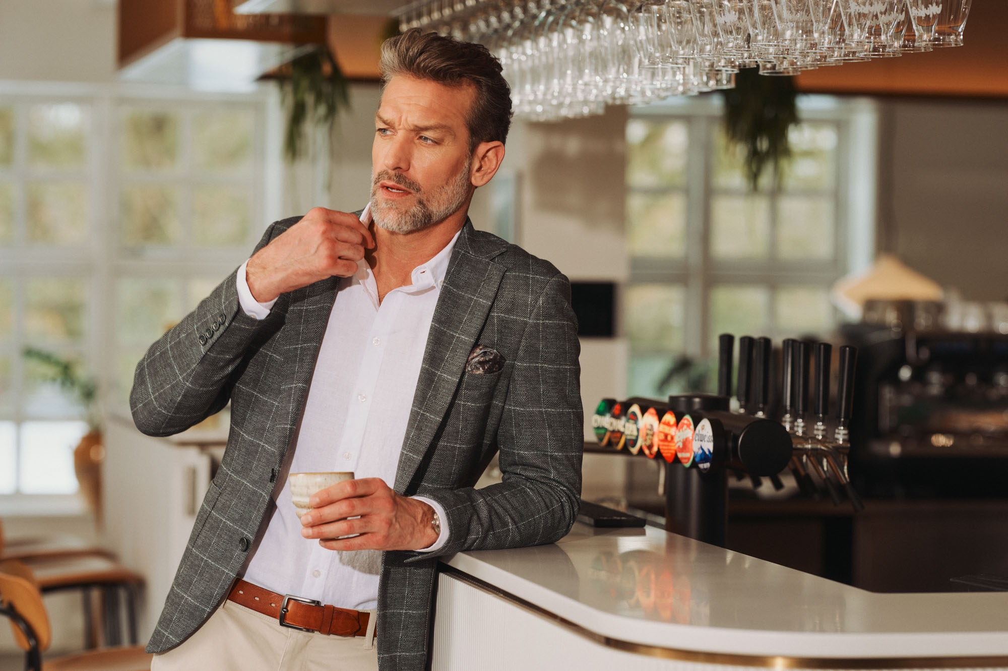 Man in plaid blazer, white shirt, adjusting collar, holding cup at a bar.