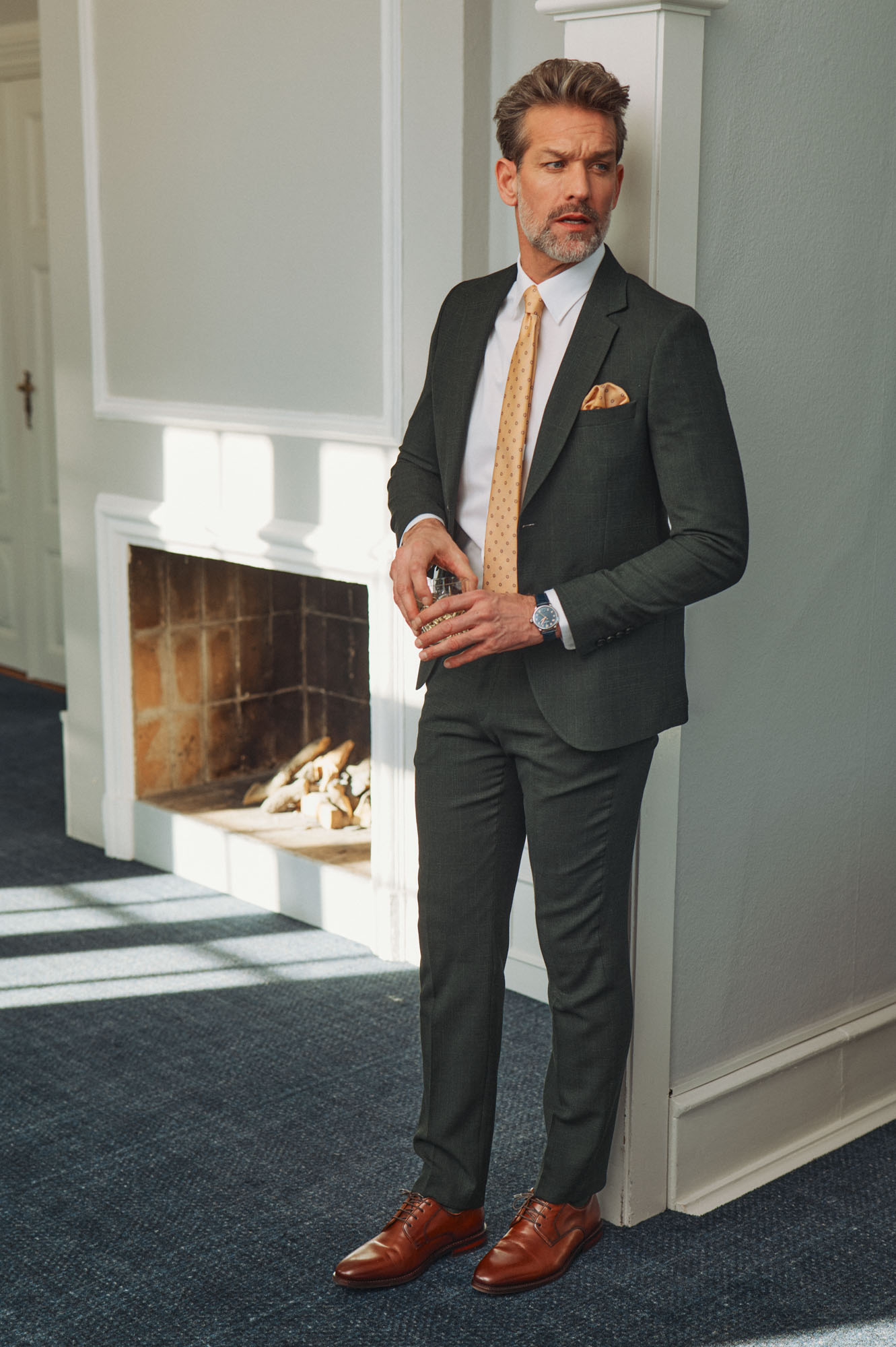 Elegant man in a green suit and yellow tie holding a glass next to a fireplace.