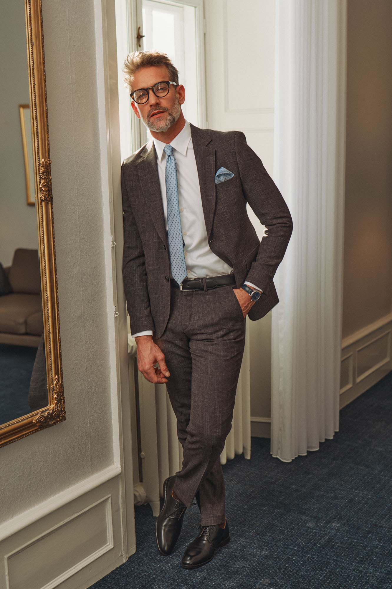 Elegant man in a brown suit, blue tie, and glasses leaning by a gold mirror indoors.