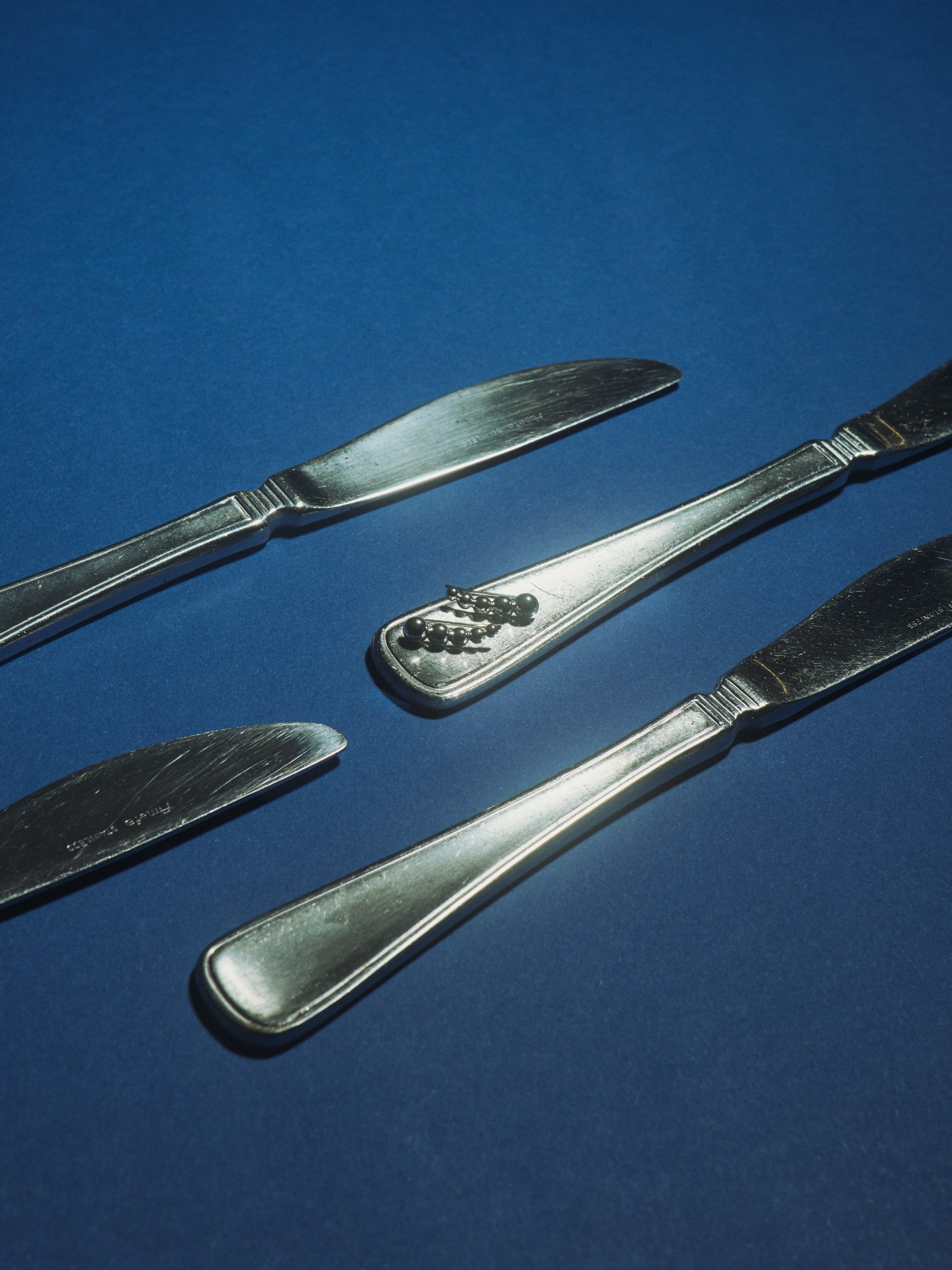 Four silver butter knives, one with small black pins, on a textured blue background.